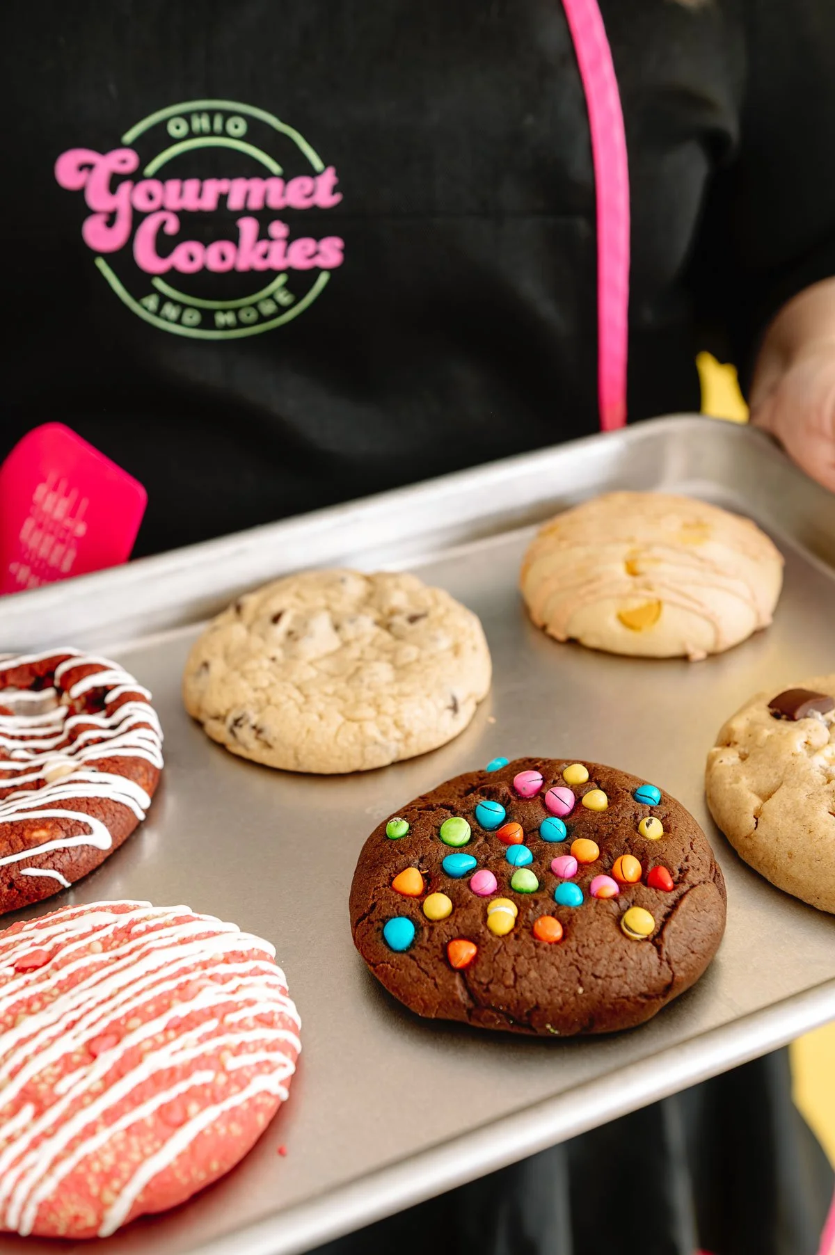 Close-up of a tray of assorted cookies, including a chocolate cookie with colorful sprinkles, a chocolate chip cookie, a white chocolate chip cookie, and two cookies with icing, held by a person wearing a black apron with pink and green text.