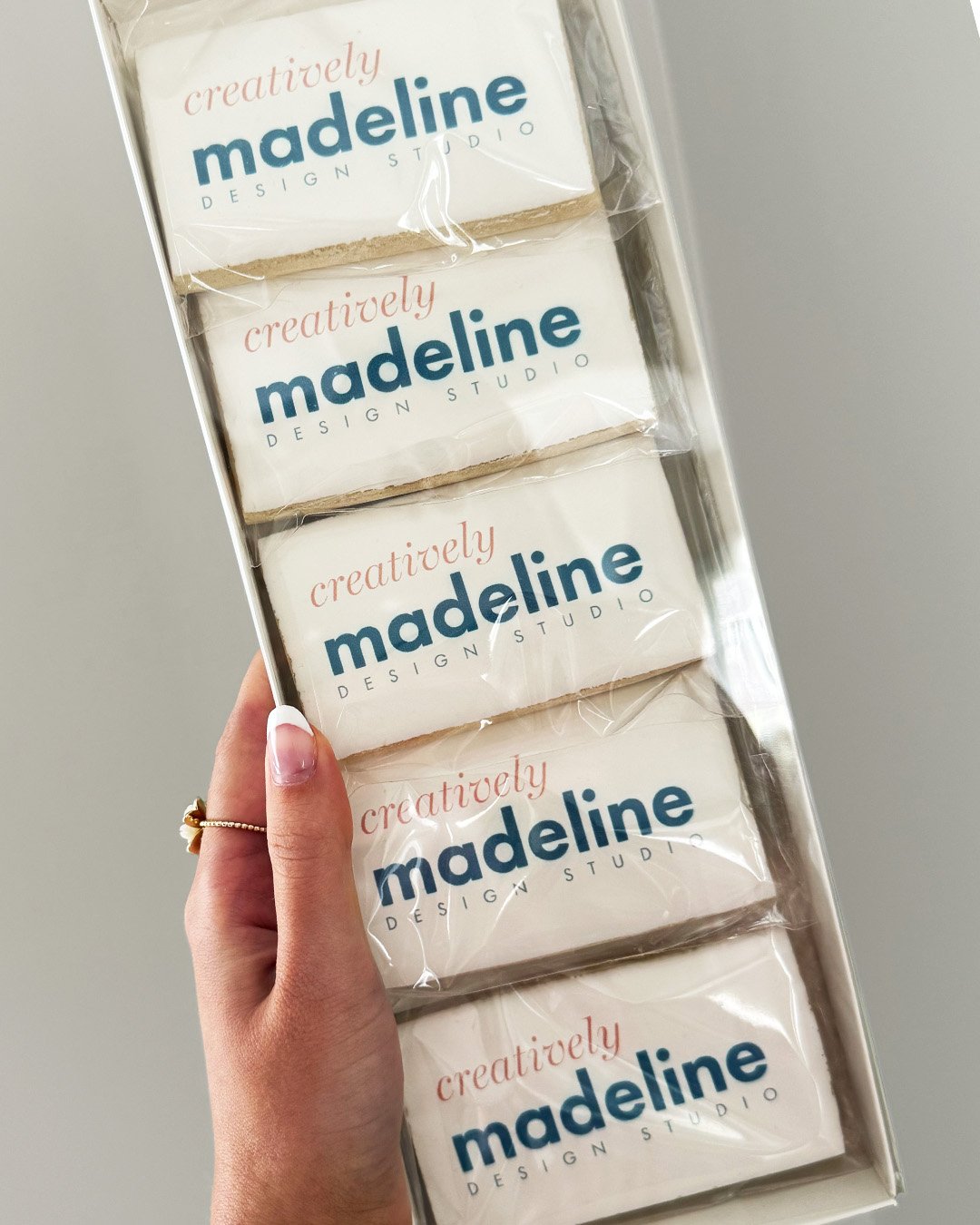A hand holding a box with five custom logo, sugar cookies wrapped in clear plastic. Each cookie has the logo "Creatively Madeline DESIGN STUDIO" printed on it in blue and pink lettering.