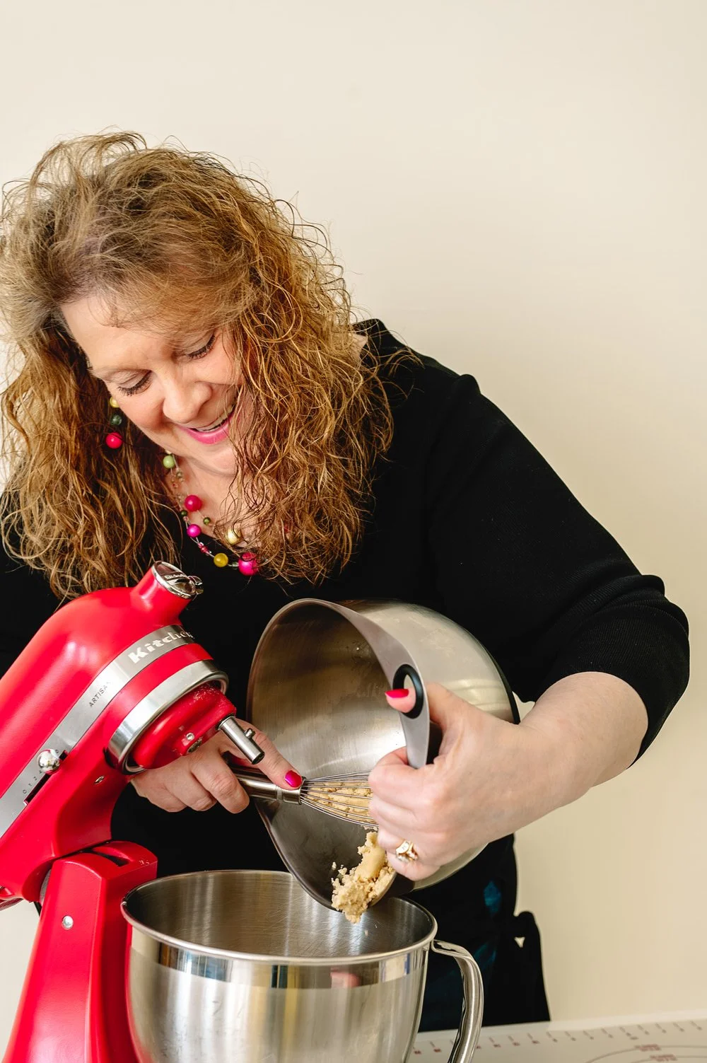 A woman with curly hair, wearing a black top and colorful jewelry, is adding cookie dough to a stand mixer in a kitchen setting.
