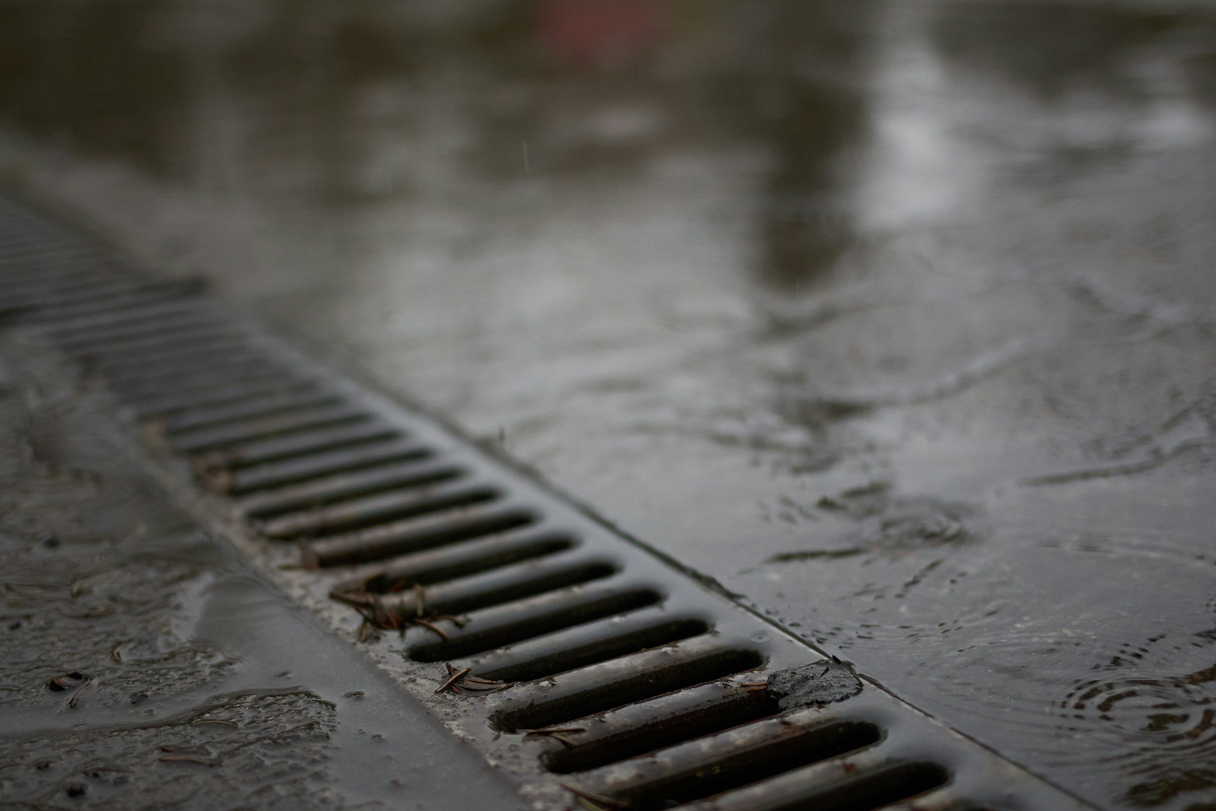 Close-up of water flowing into a drain on a wet pavement with raindrops creating ripples.