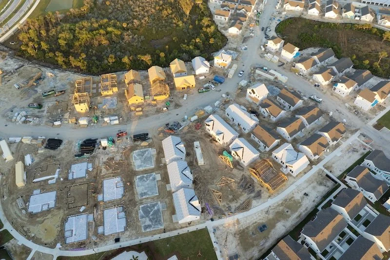 Aerial view of a residential construction site with multiple houses in various stages of being built, some with framing and others with roofing, surrounded by completed houses and streets.