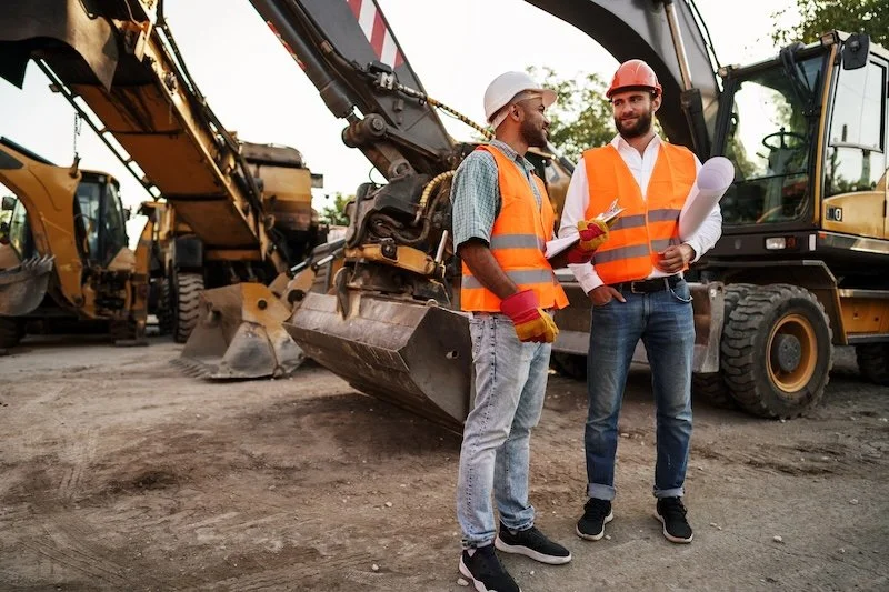 Two construction workers in orange safety vests and helmets talking in front of large construction equipment at a site.