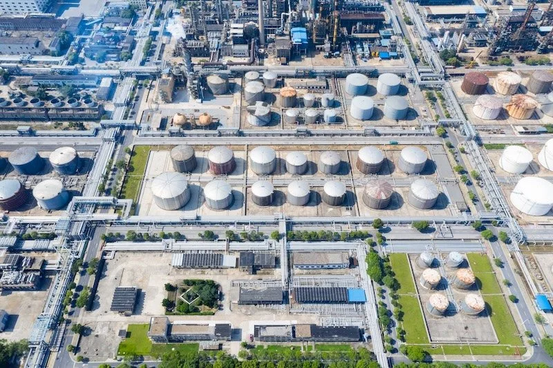 Aerial view of an industrial plant with multiple storage tanks, pipelines, and infrastructure, including greenery and buildings.