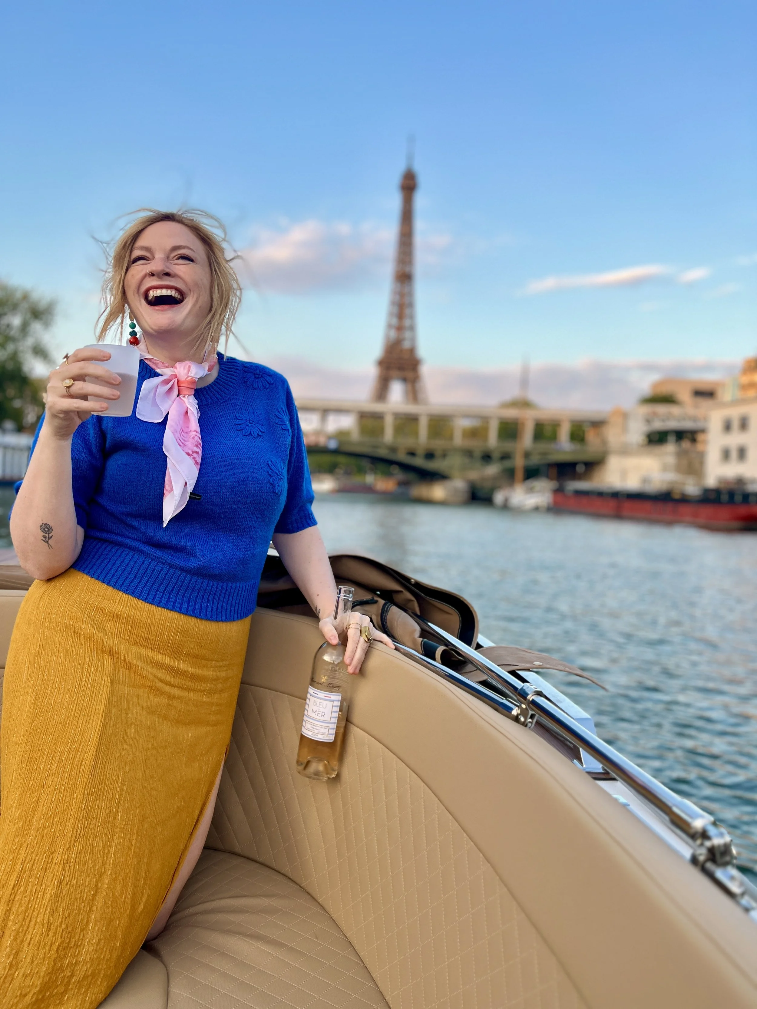 Woman laughing on a boat with a view of the Eiffel Tower in the background, holding a drink and standing next to a bottle.