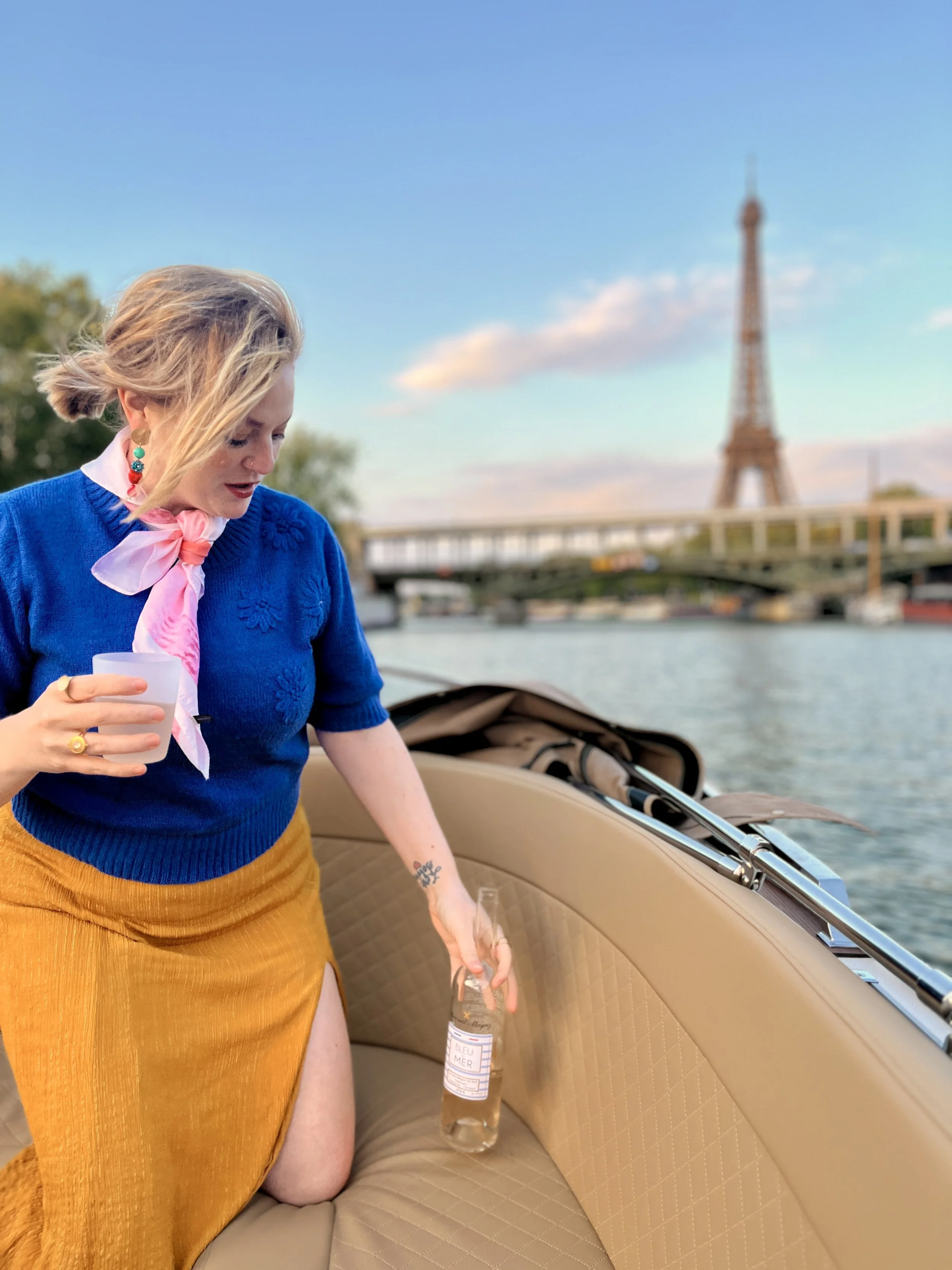 A woman with blonde hair, wearing a blue sweater, a pink scarf, and an orange skirt, kneeling on a boat with a bottle and a glass. The Eiffel Tower is visible in the background over the river.