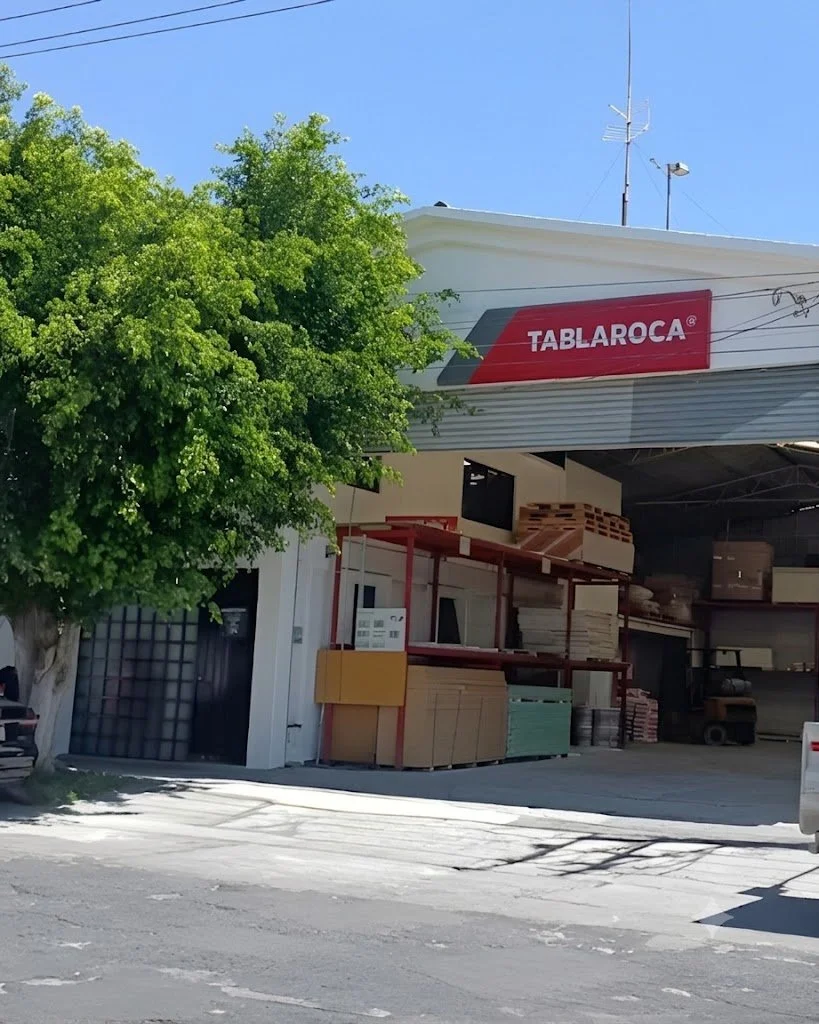 Vista de una tienda llamada TABLAROCA que vende materiales de construcción, con estanterías de madera y cartón, y un árbol grande en la acera.