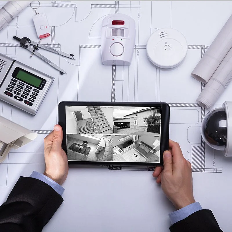 Person holding a tablet showing security camera footage on architectural blueprint table with tools and security devices.