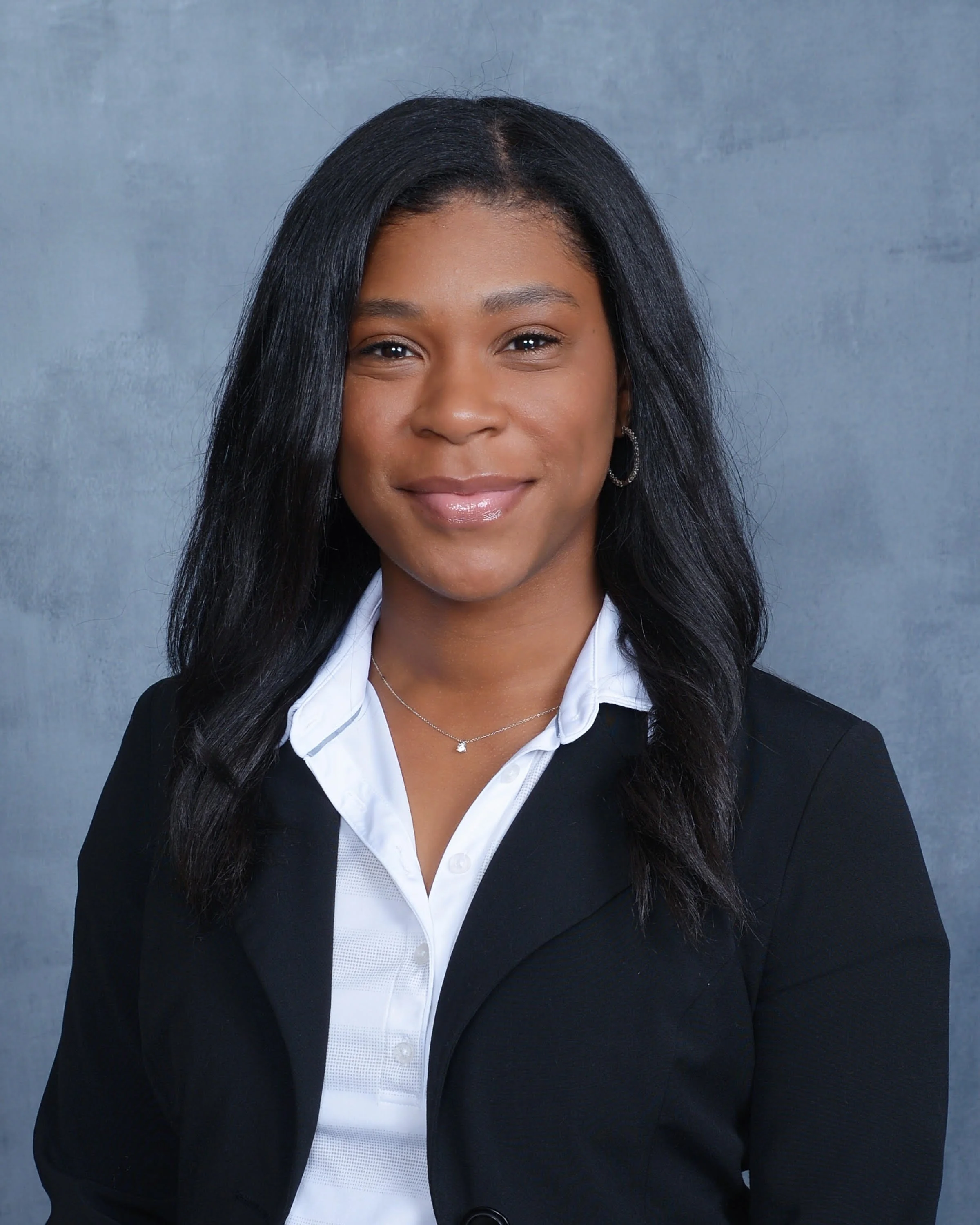 Close-up of a woman with long black hair, smiling, wearing a white collared shirt and a black blazer, with a silver necklace and hoop earrings, against a gray background.