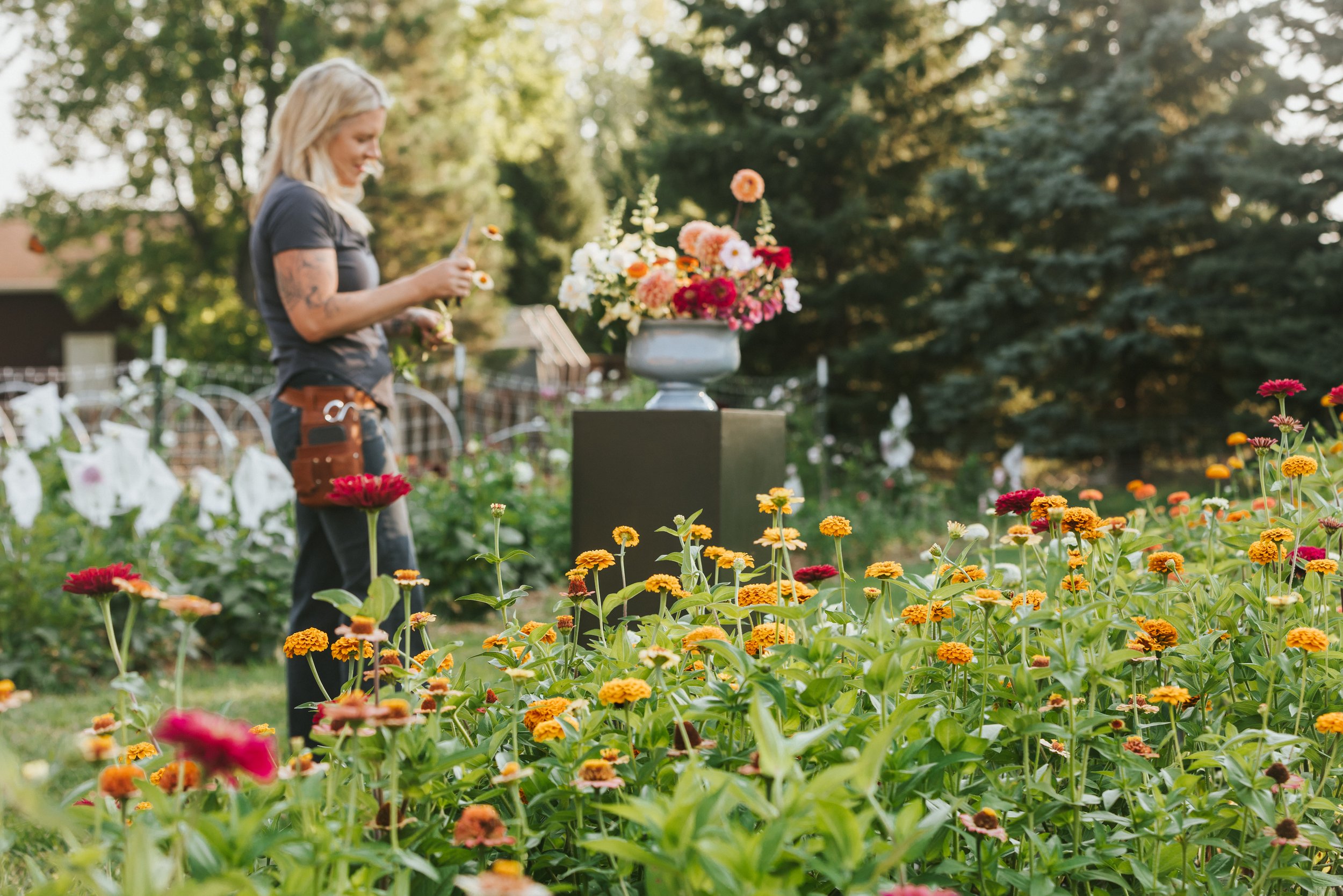 A woman with blonde hair and tattoos on her arms tending to a garden of colorful flowers, with a large floral arrangement on a pedestal in the background, surrounded by lush green trees.