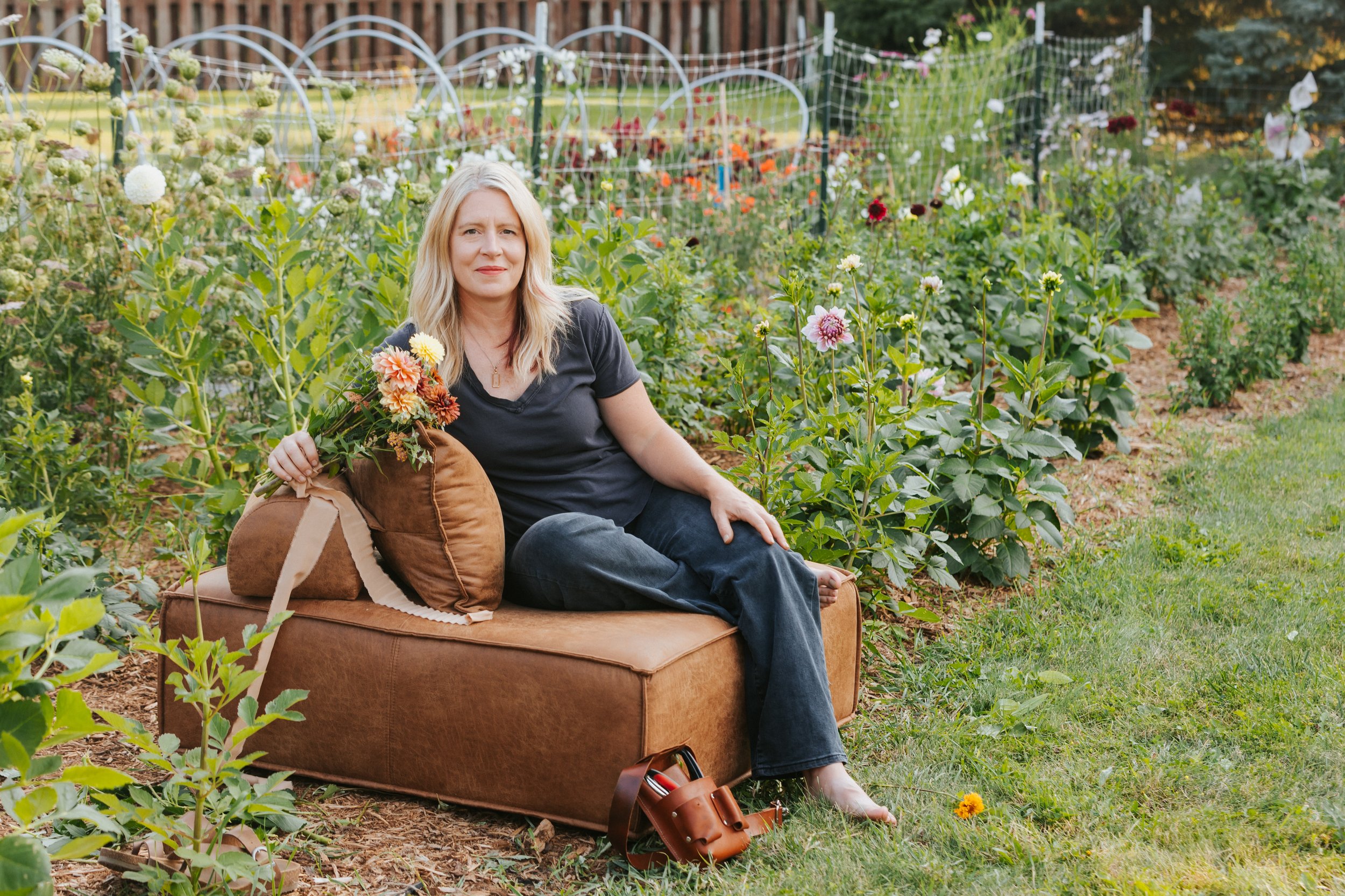 A woman sitting on a brown sofa in a flower garden, holding a bouquet of flowers, with colorful blooms and green plants in the background.