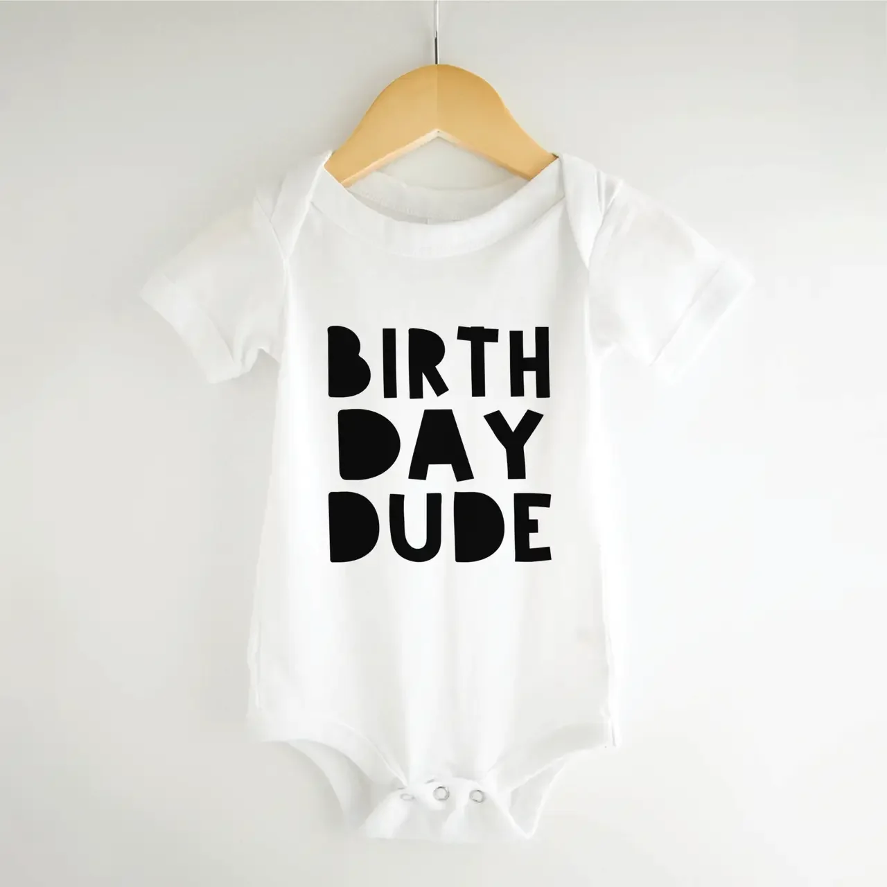 A white "Birthday Dude" infant onesie on a wooden hanger, a perfect gift or outfit found in our birthday party supplies for Madrona and Madison Park families.