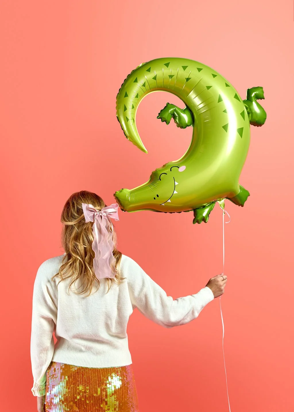 A whimsical green alligator birthday balloon being held against a coral backdrop, a fun example of the curated party supplies serving families in Mount Baker and the Central District.