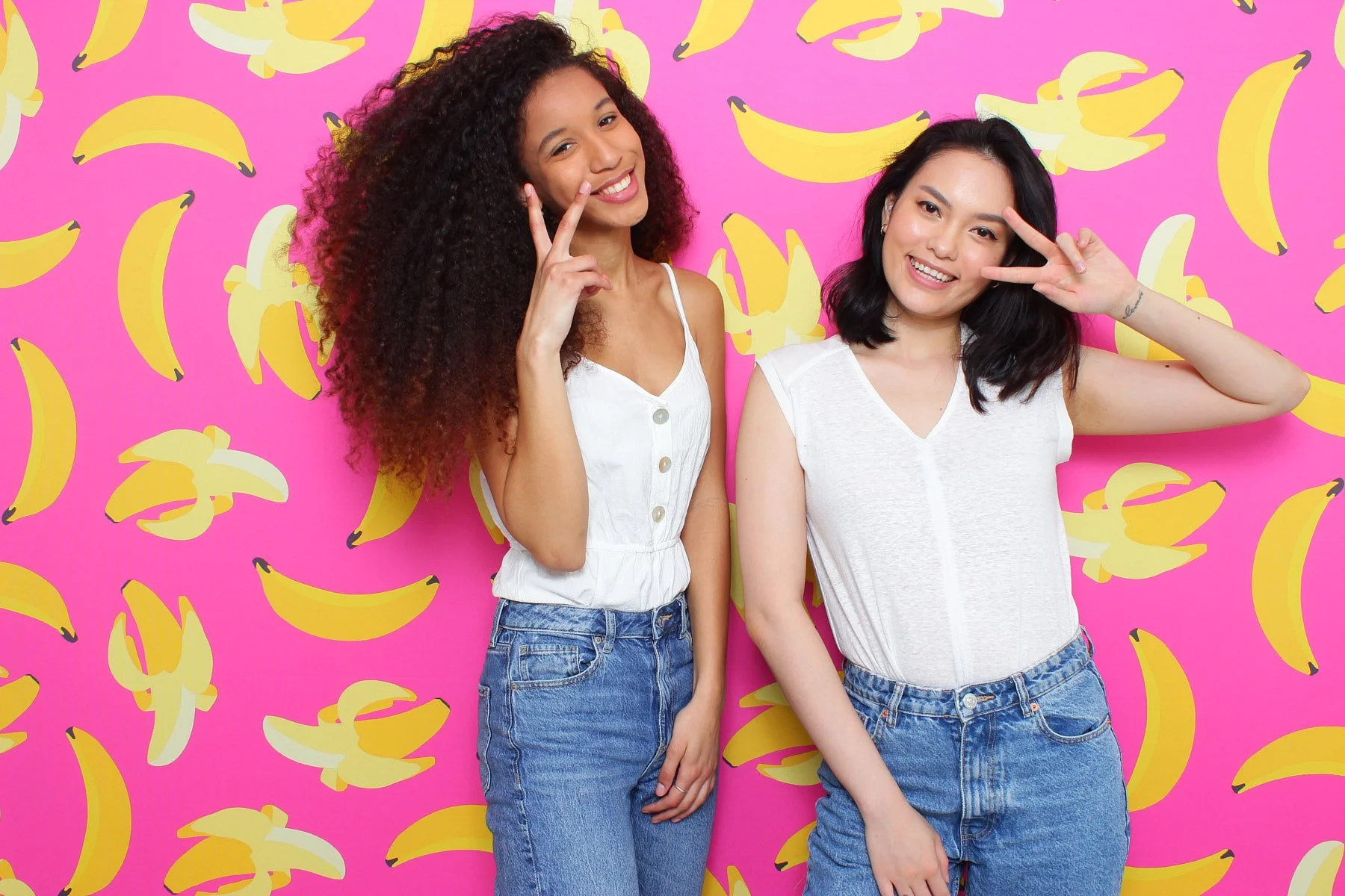 Two friends posing with peace signs in front of a vibrant yellow banana print wall, a fun example of the curated party supplies and photo backdrops available for Seattle celebrations in the Central District and Leschi.