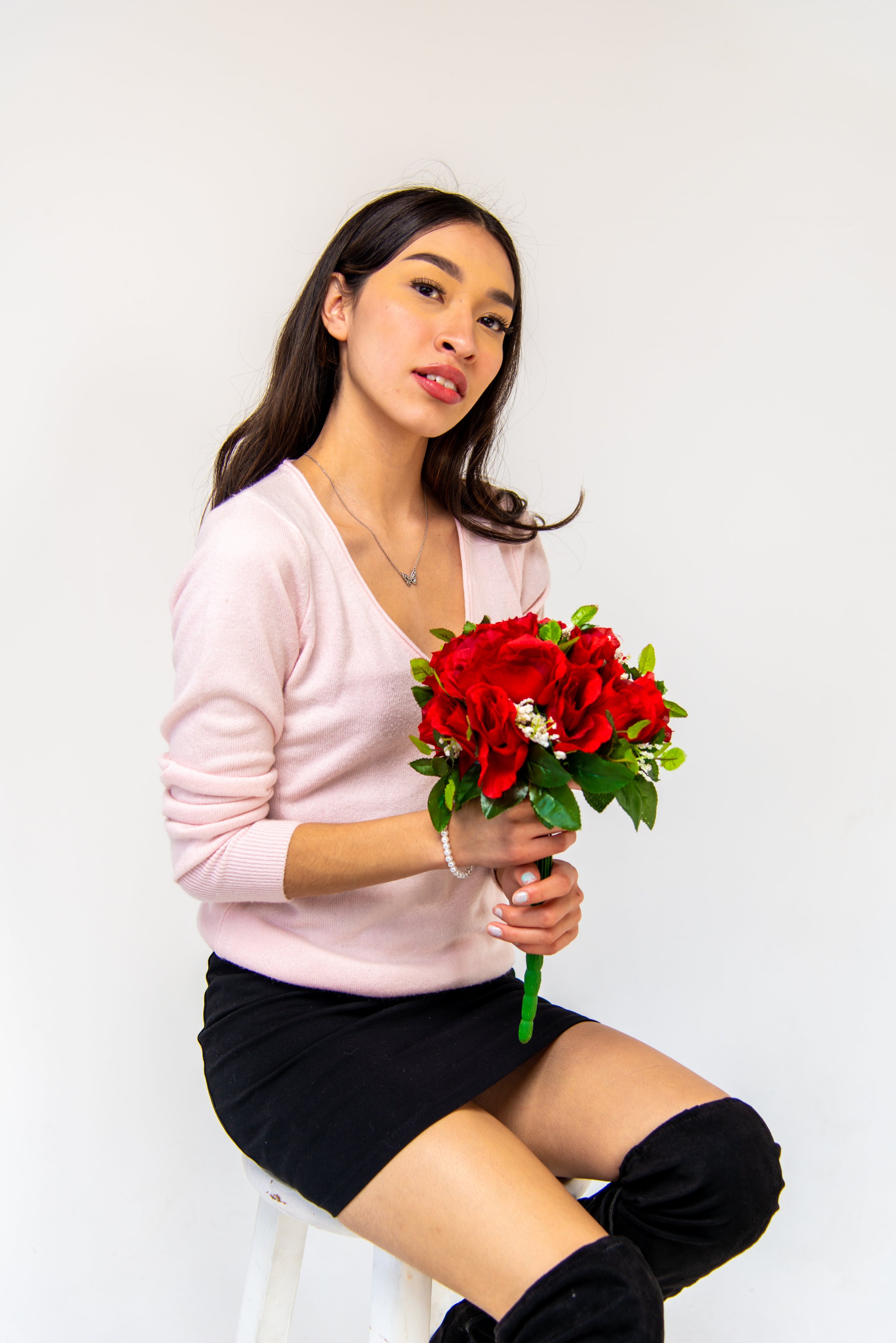 A woman with long dark hair, wearing a pink sweater and black skirt, is sitting on a white stool holding a bouquet of red roses with green leaves, against a plain white background.