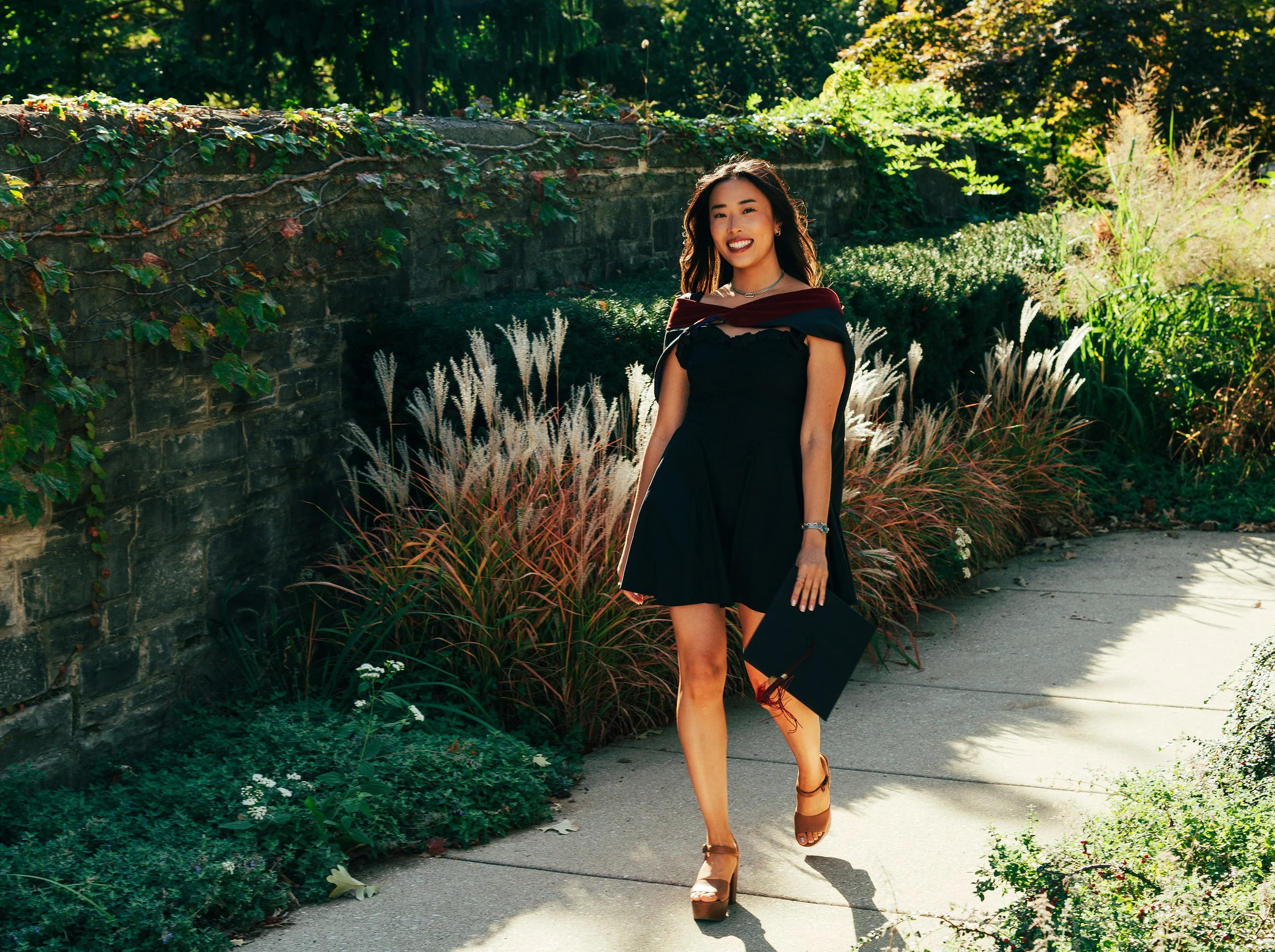 A woman in a black dress, smiling and walking on a sidewalk in a garden area with a brick wall and lush green plants.
