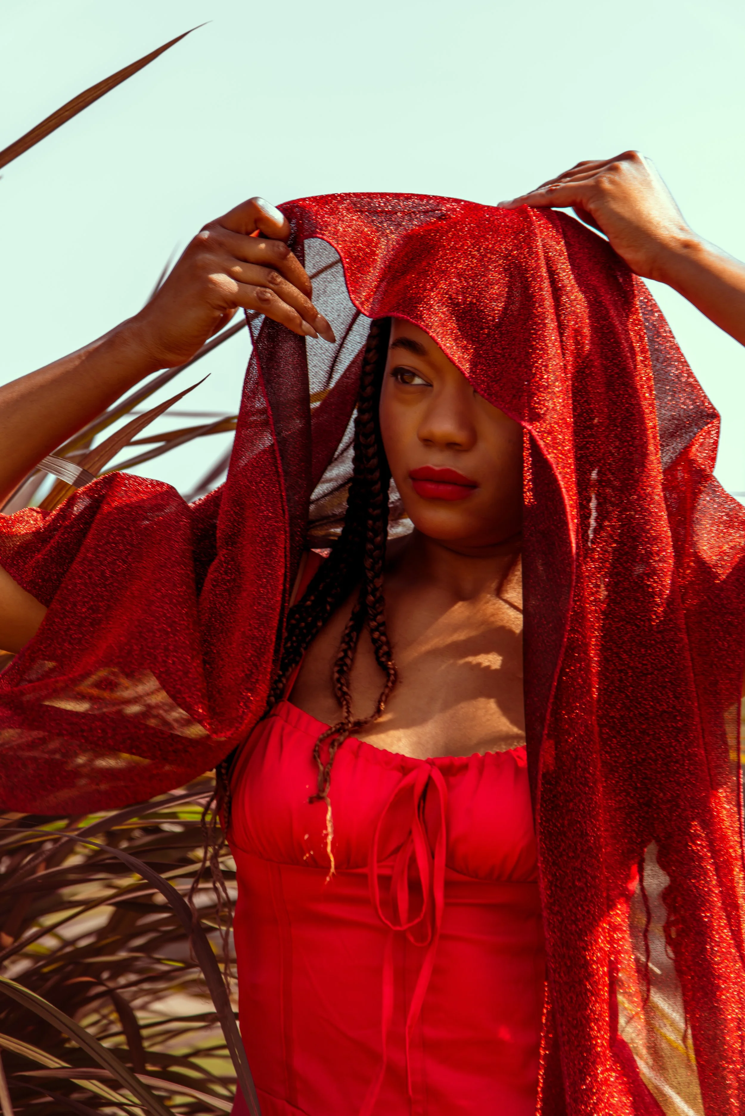 A woman with braided hair is adjusting a red, shimmery, semi-transparent fabric over her head outdoors, with tall grass or plants and a clear sky in the background.