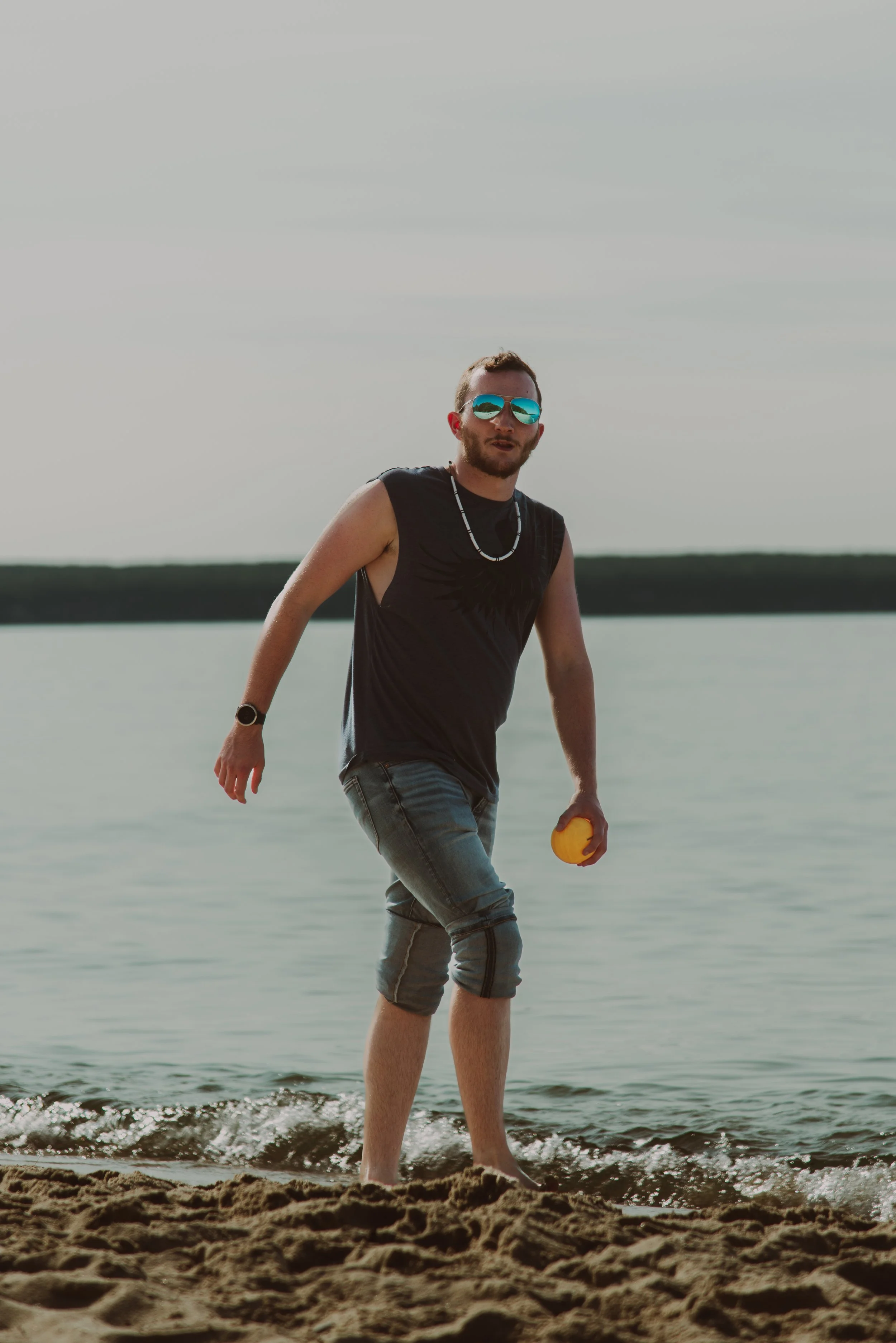 A man with a beard wearing reflective sunglasses, a black sleeveless shirt, and gray shorts, walking on a beach and holding a yellow ball.