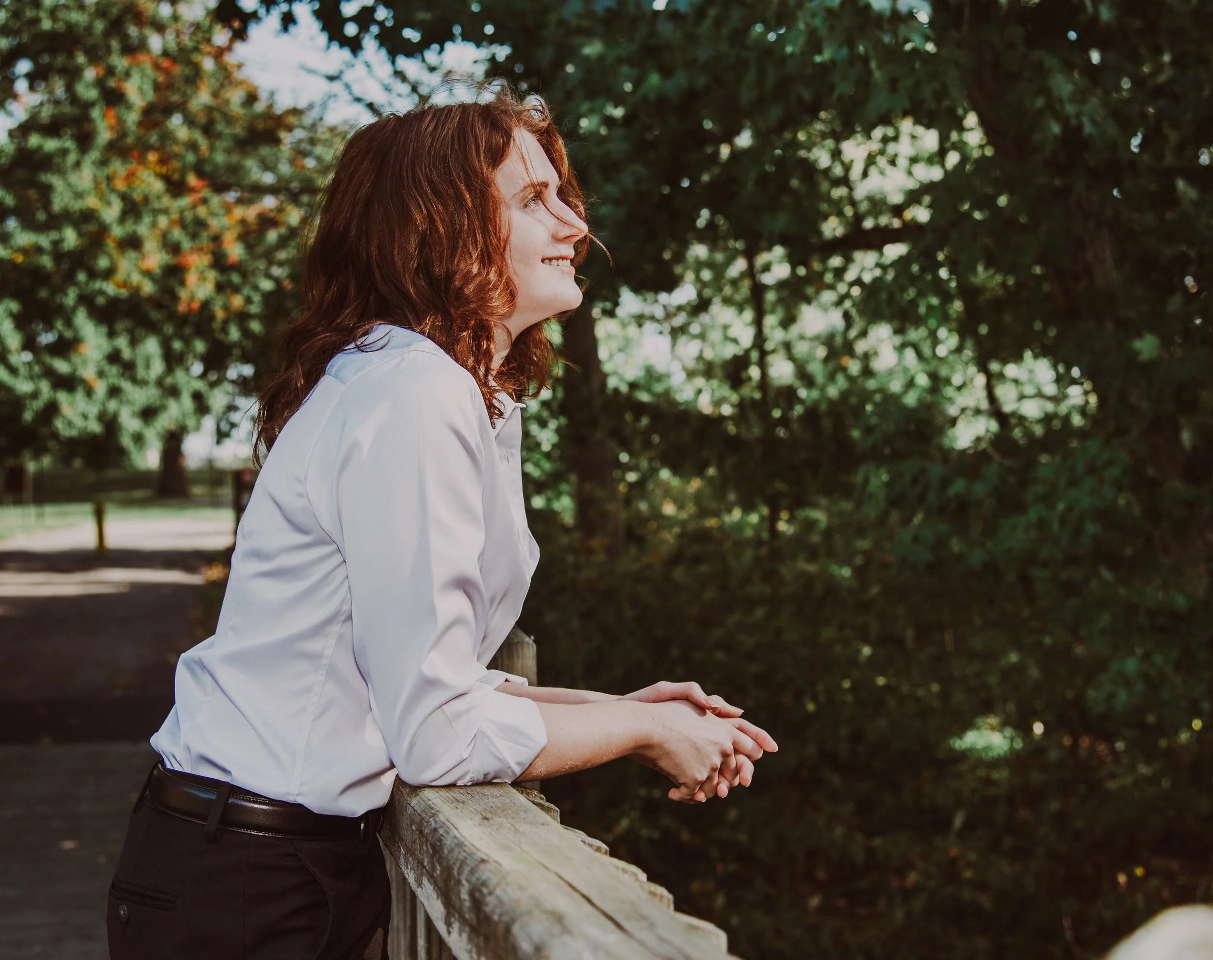 A woman with curly red hair wearing a white shirt sitting on a park bench, smiling and looking to the right, with green trees in the background.