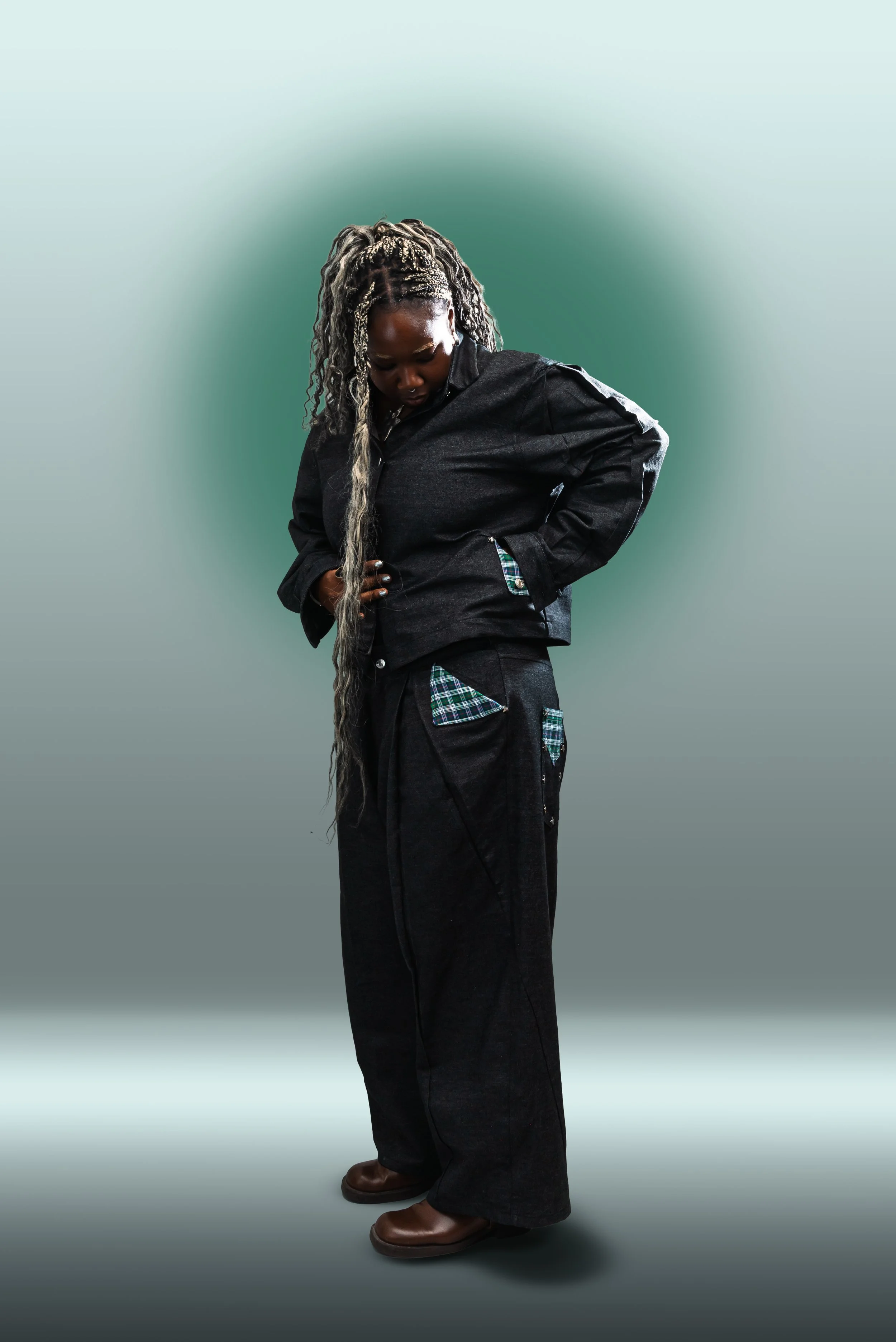 A person with long, braided, blonde and black hair, wearing a black jacket and black pants, looking down with one hand on their waist, standing against a gradient background.