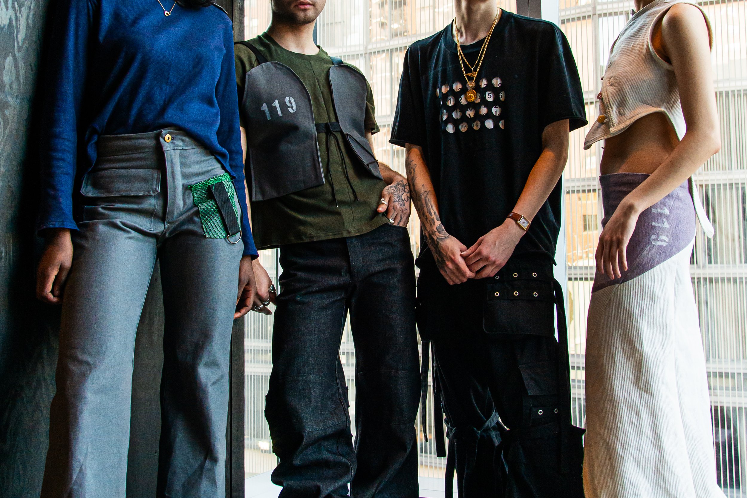 Close-up of four individuals standing together in casual streetwear, showing their torsos and arms, in front of a glass window with building reflections.