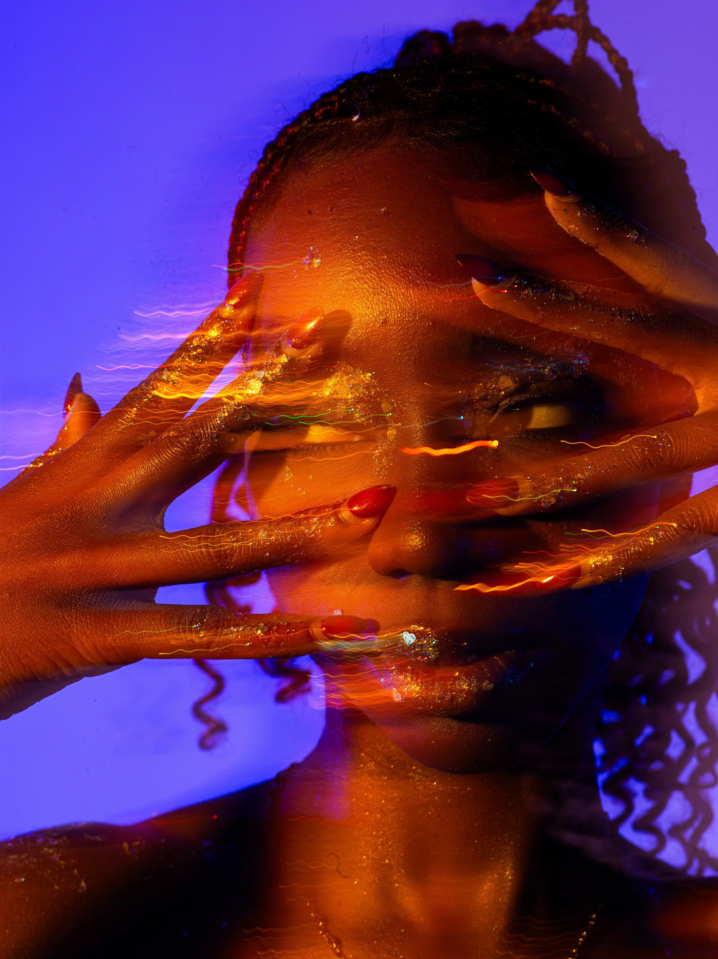 A person with dark skin and curly hair holding their face with hands painted with metallic nail polish, illuminated by vibrant orange and blue lighting, creating a dreamy, abstract effect.