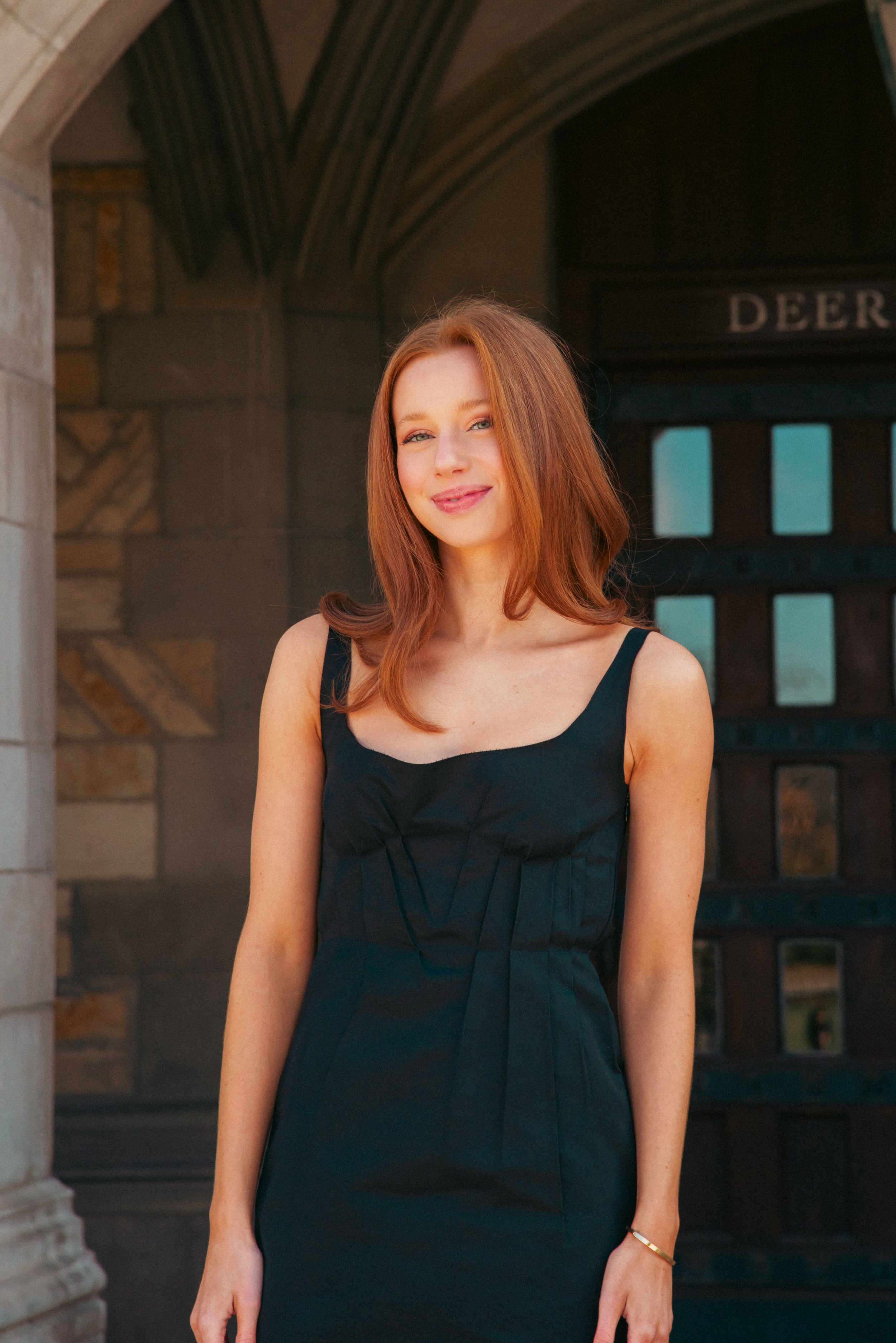 A young woman with red hair and fair skin wearing a sleeveless black dress, standing outdoors in front of a building with stone archways and a dark wooden door.