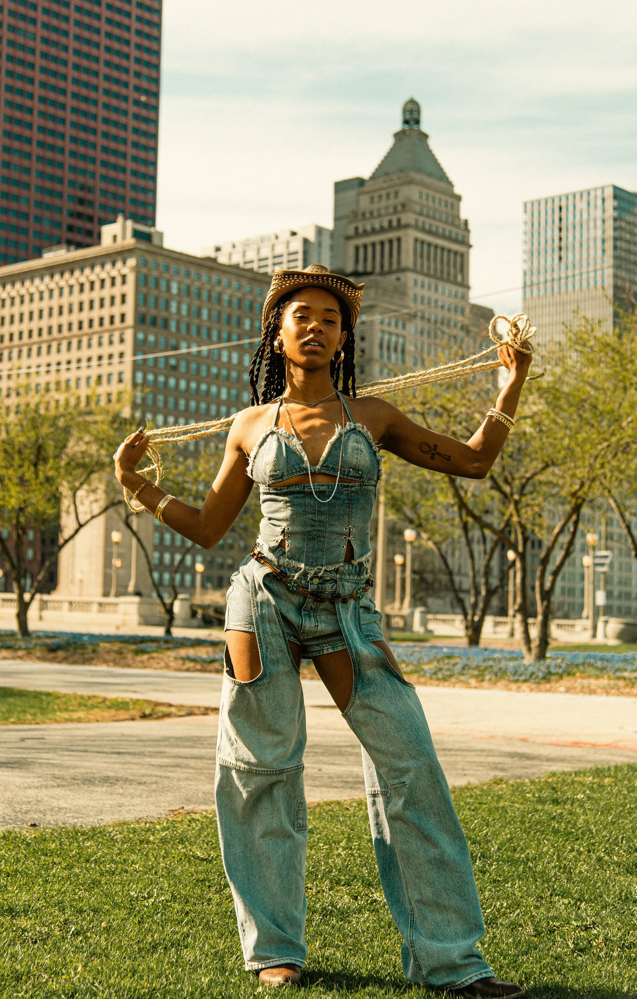 Young woman with dreadlocks wearing a cowboy hat, denim crop top, and baggy jeans holding a rope with a cityscape in the background.