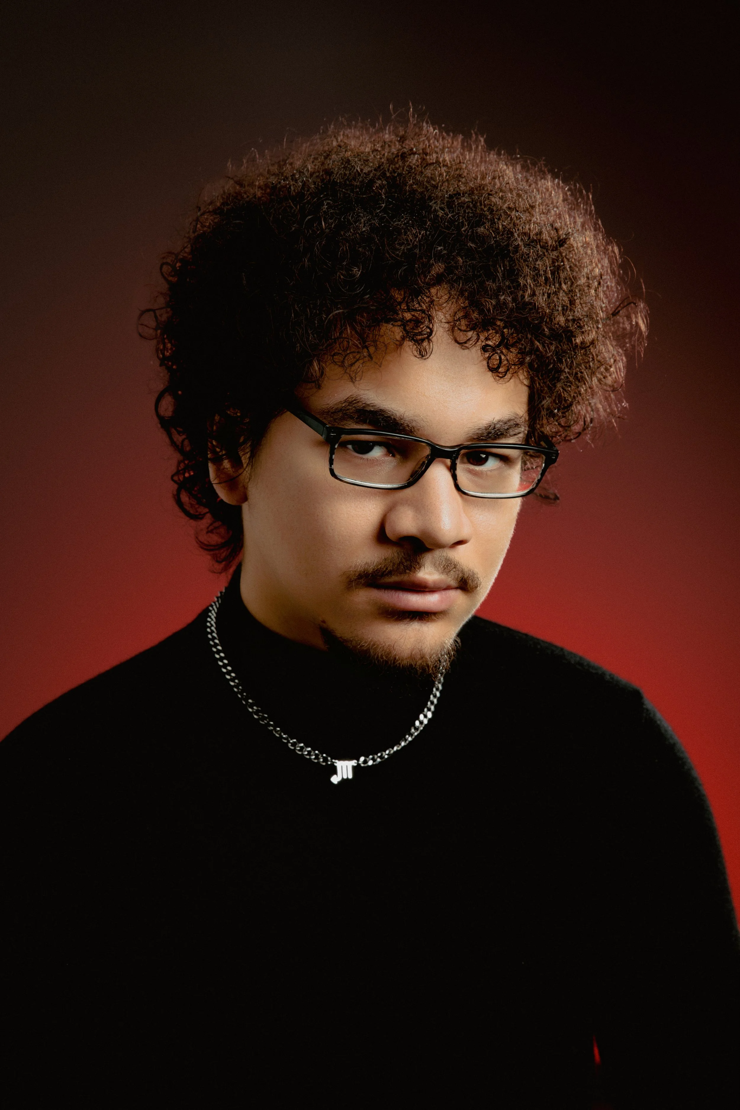A man with curly hair, glasses, a goatee, wearing a black turtleneck and a silver necklace with initials, against a red background.