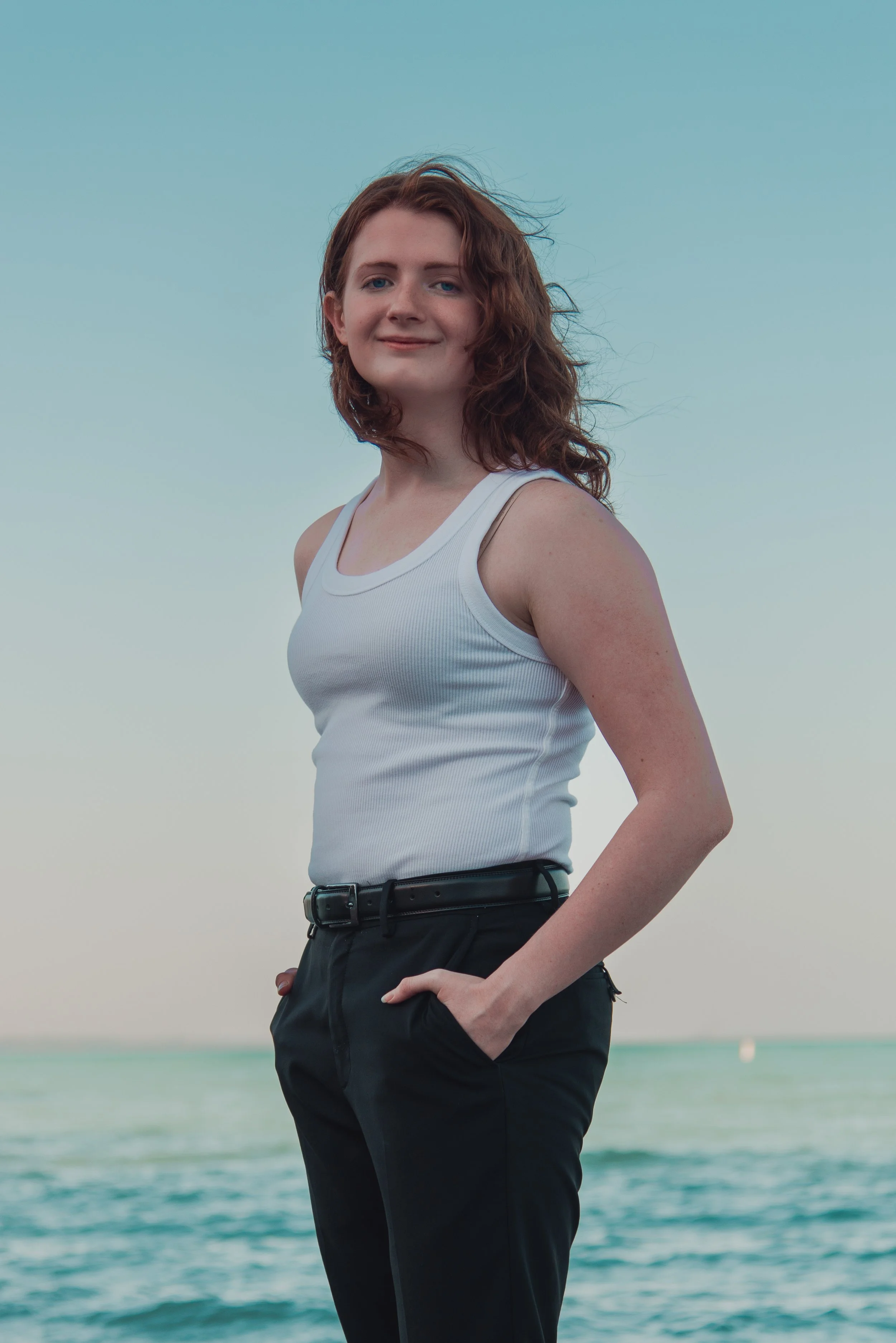 A woman with wavy brown hair wearing a white tank top and black pants standing on a beach with ocean waves in the background.