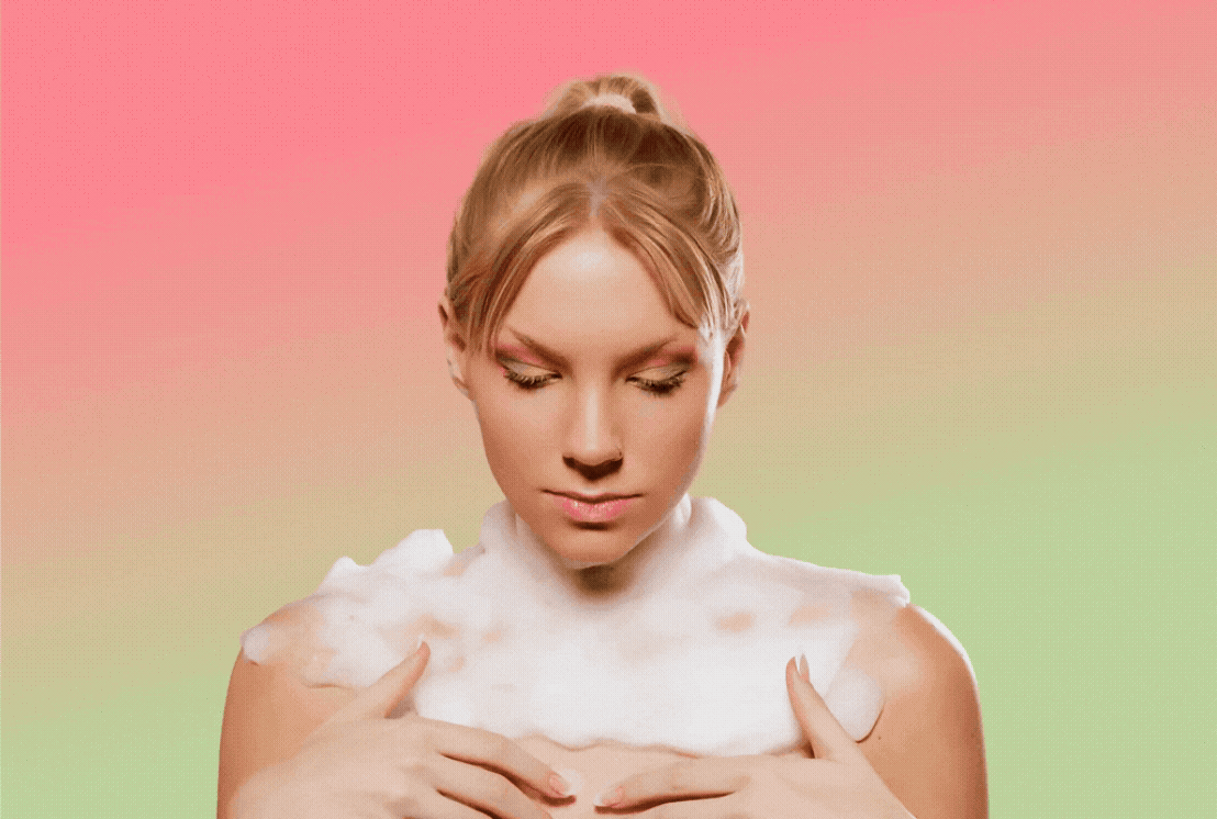 A woman with blonde hair in a bun, eyes closed, and wearing white foam on her shoulders, against a gradient pink and green background.