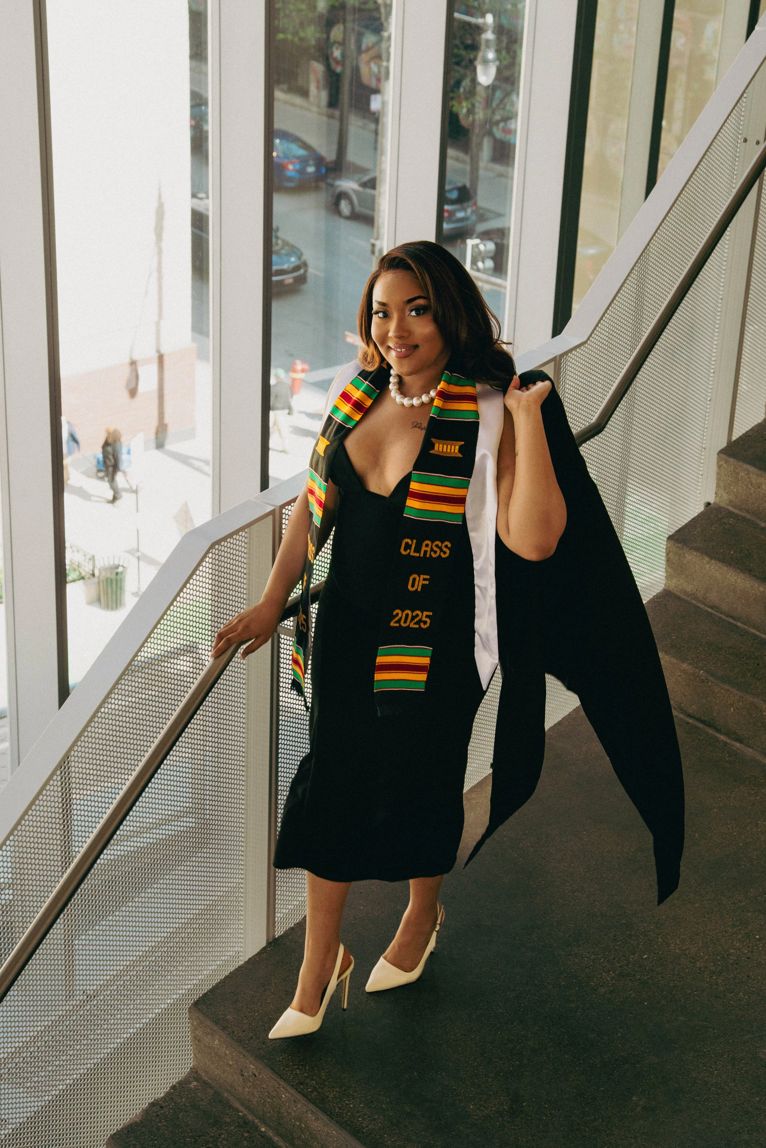 A young woman in a black dress and white high heels standing on stairs inside a building, wearing a graduation stole that says 'Class of 2025' and a white pearl necklace, with a large window behind her showing a street scene.