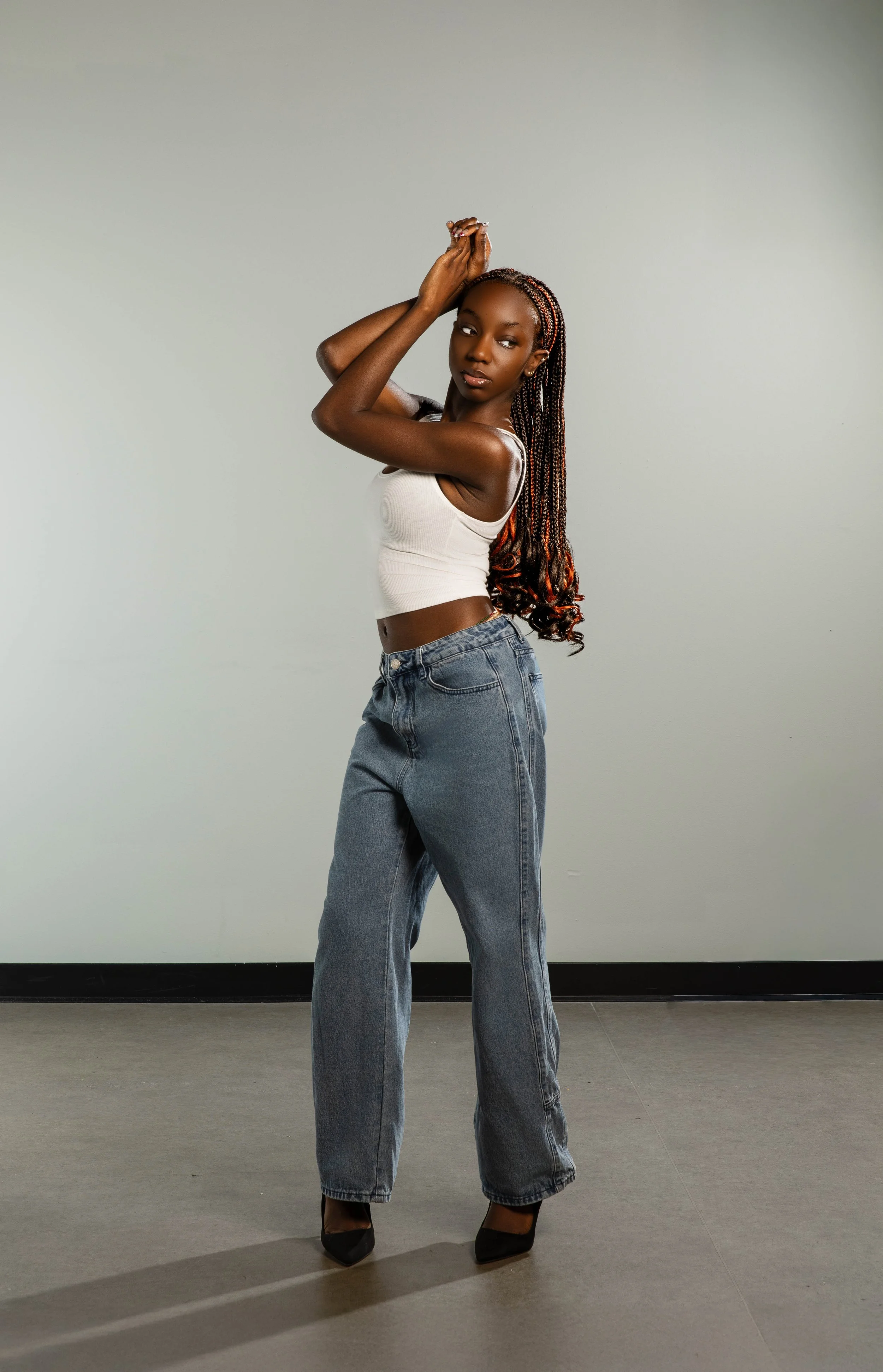 A young woman with braided hair, wearing a white tank top and high-waisted jeans, standing against a plain light gray wall, posing with her left arm bent and her hand touching her head while she looks towards the camera.