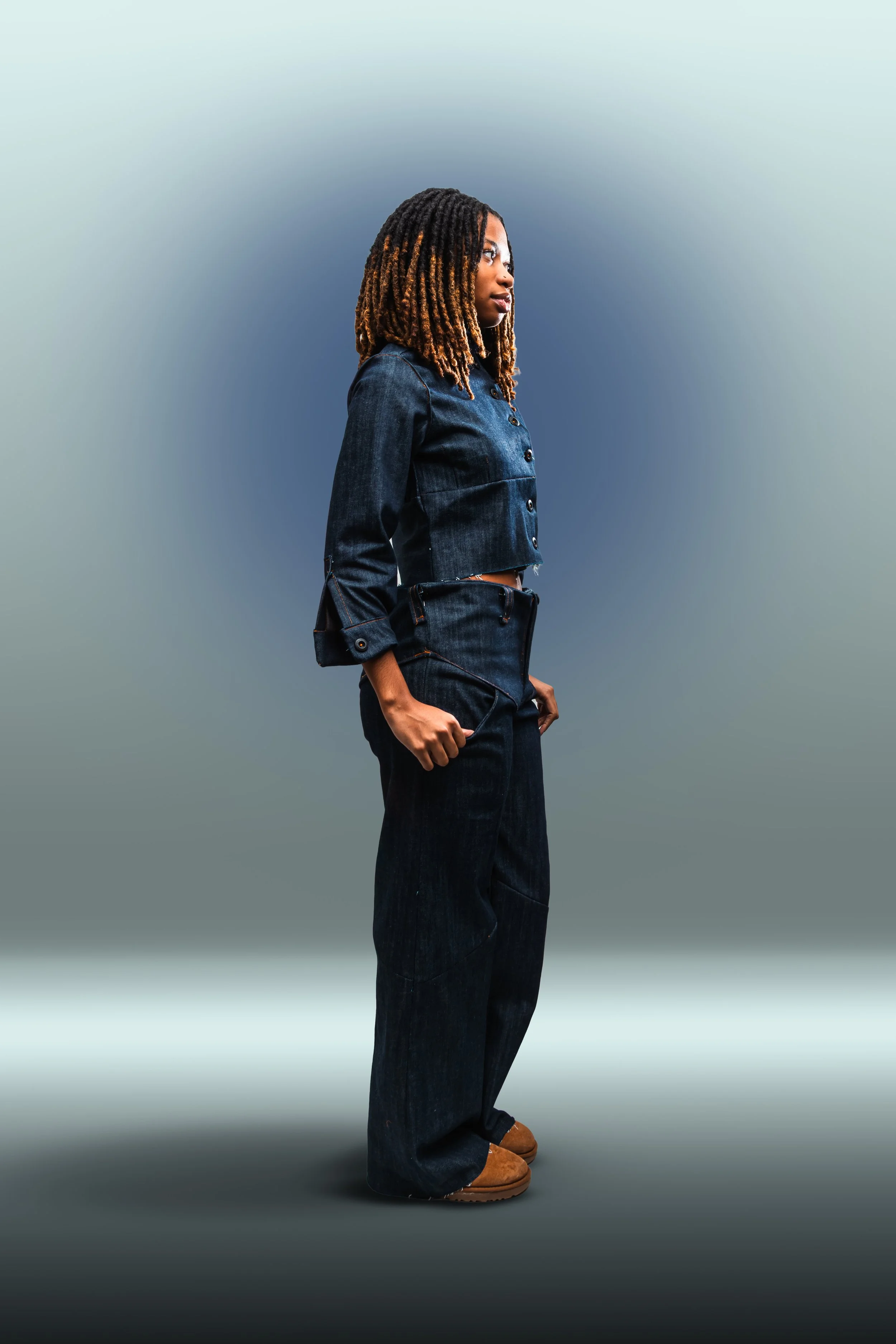 Side view of a woman with dreadlocks wearing a dark denim jacket, matching wide-leg jeans, and brown boots, standing against a gradient blue background.