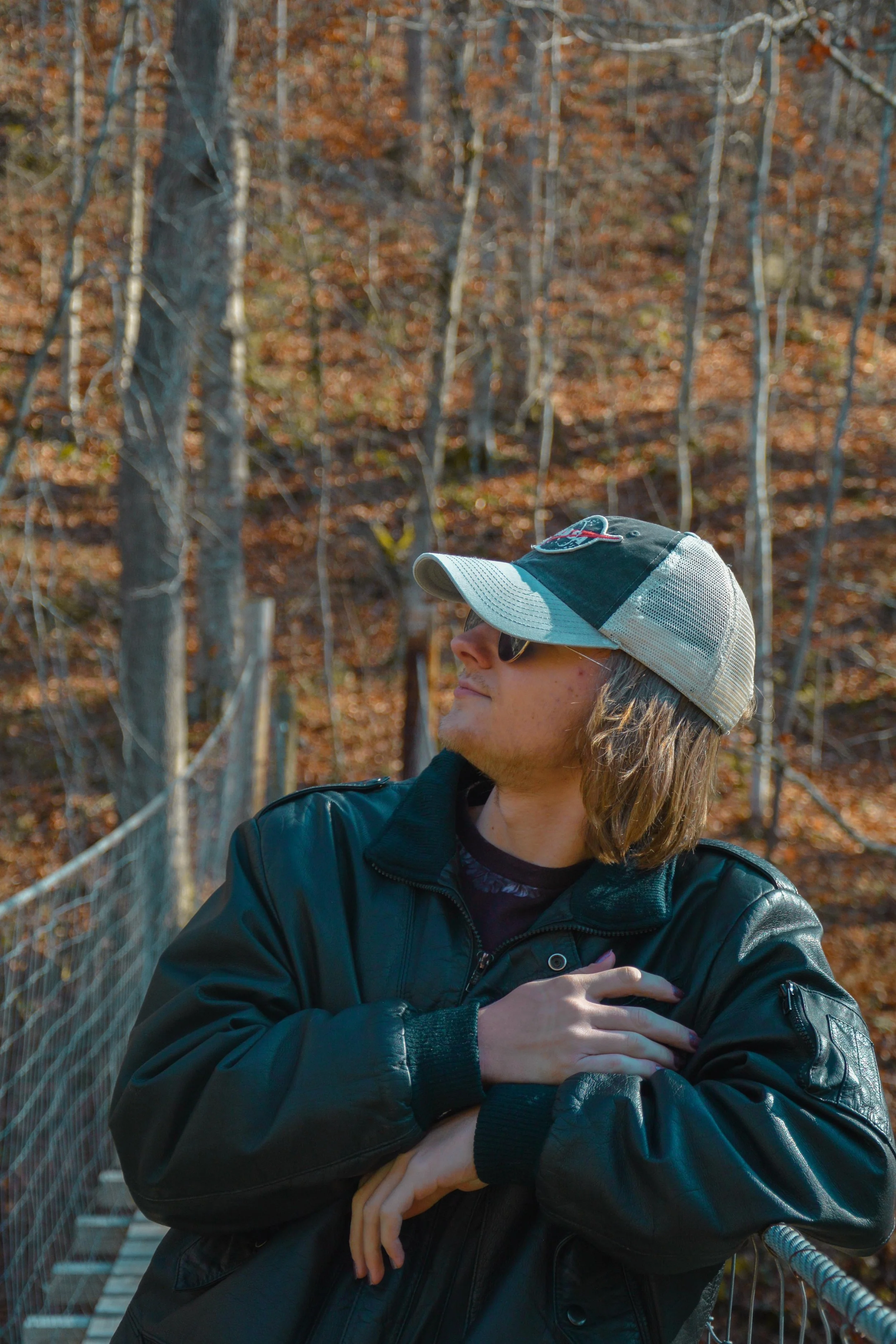 A person with long hair wearing sunglasses, a cap, and a black leather jacket, standing on a bridge in a forest with autumn foliage.