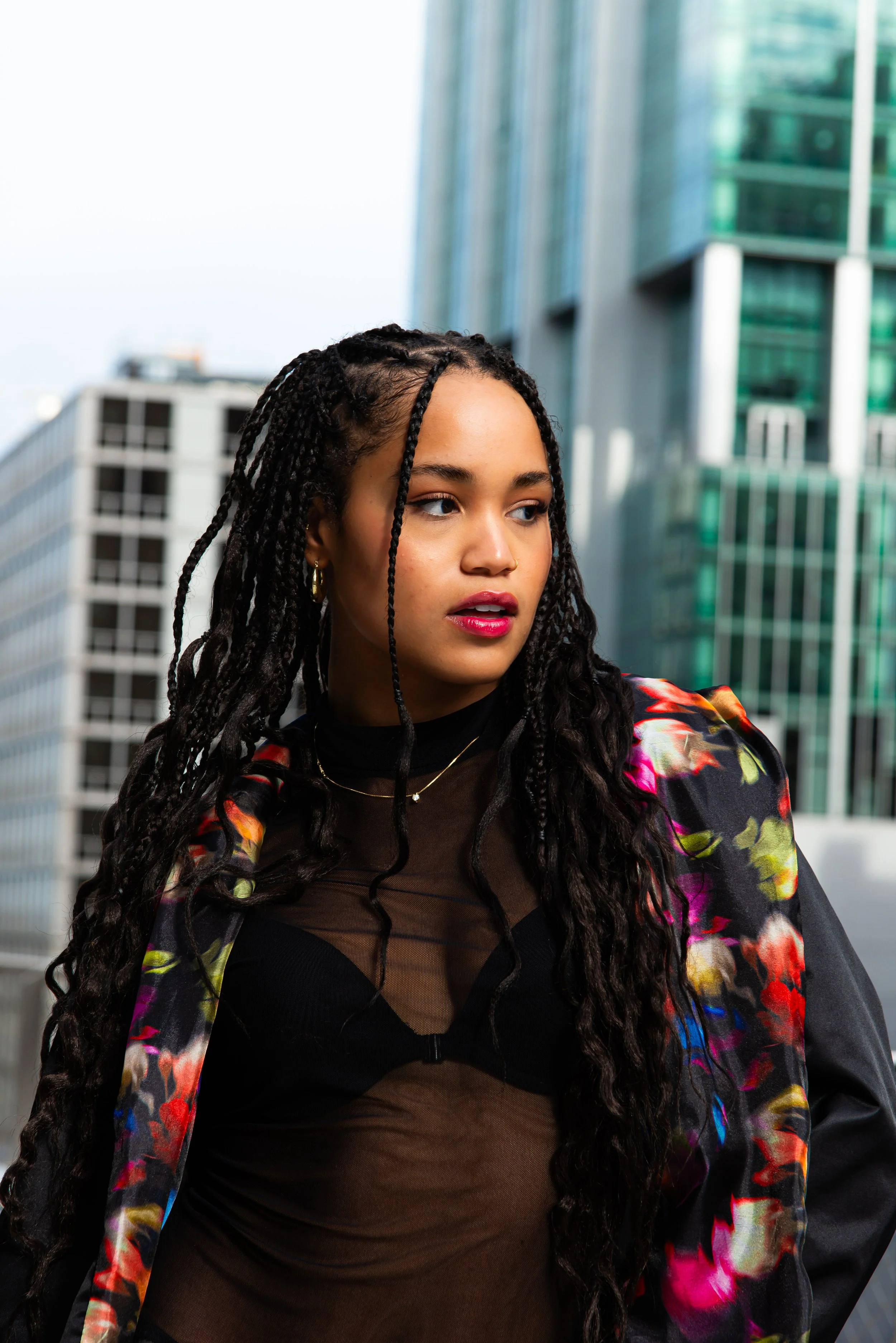 A young woman with long, curly hair styled in twists, wearing a black mesh top and a colorful floral jacket, standing outdoors in an urban area near tall glass buildings.
