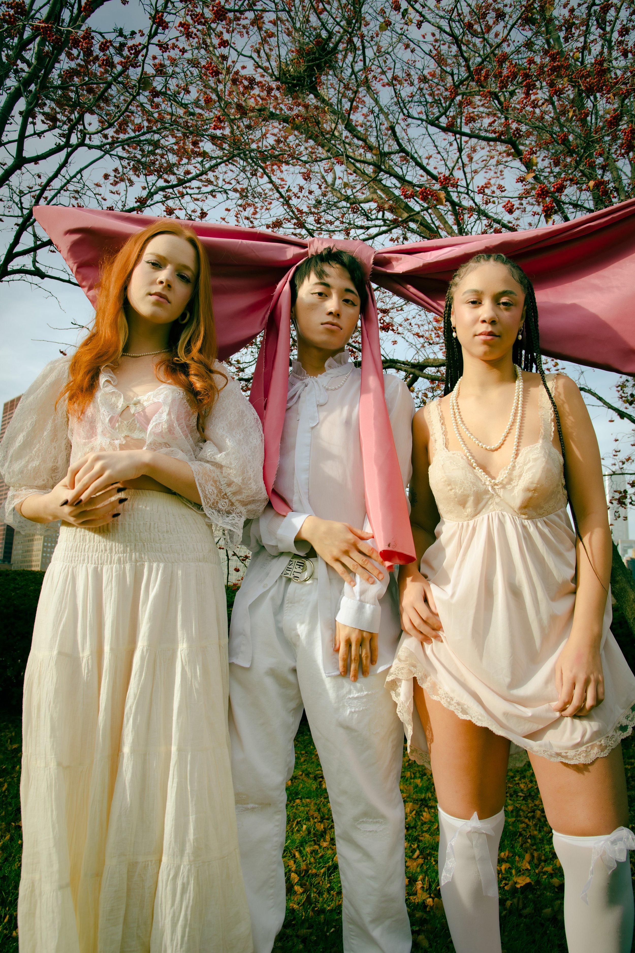 Three young women standing outdoors beneath a tree with red berries, wearing vintage-style white and cream dresses with pearls, and a pink fabric draped above their heads.