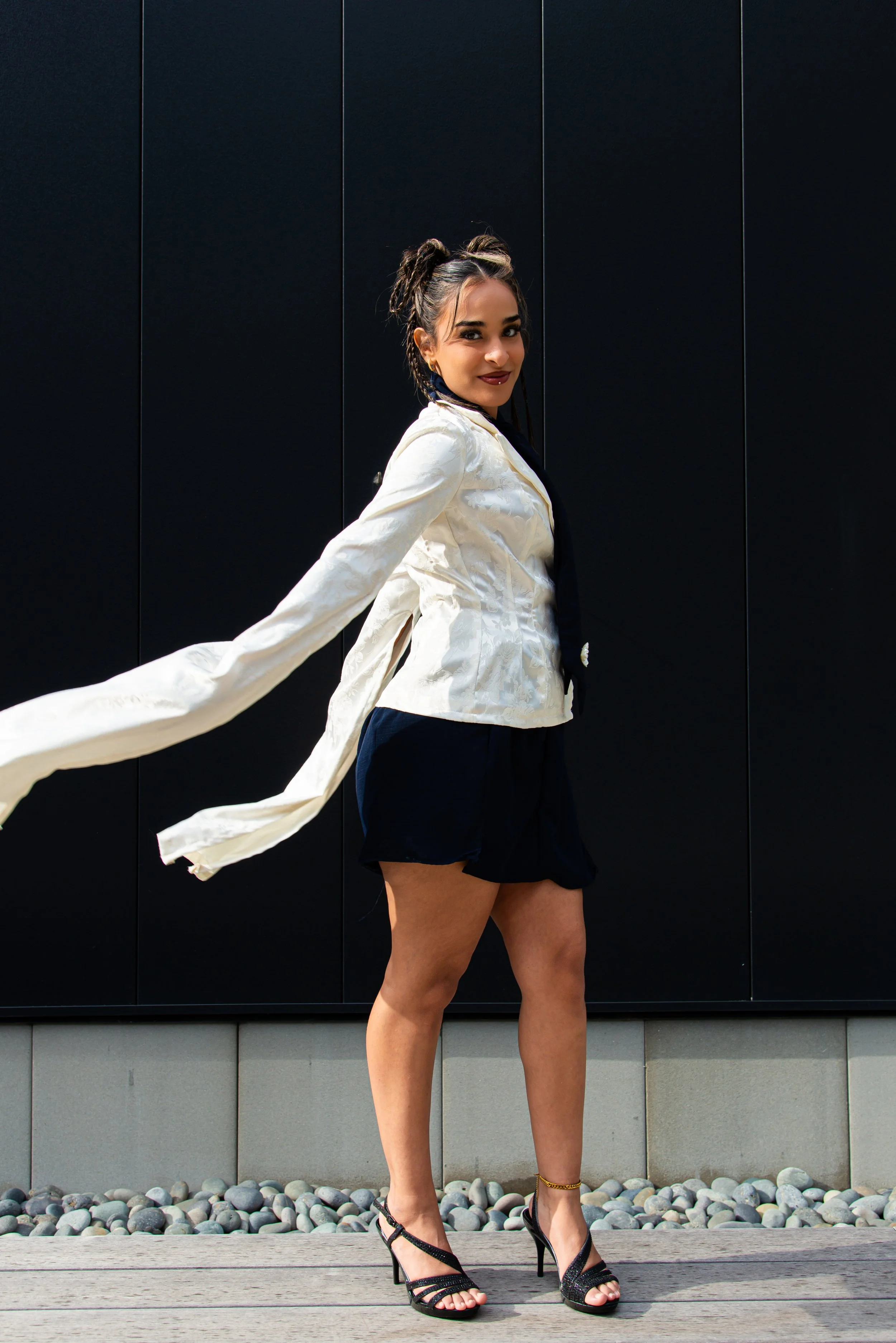 A woman in a white blazer and black shorts posing outdoors in front of a black wall, wearing black high-heeled sandals and gold jewelry.