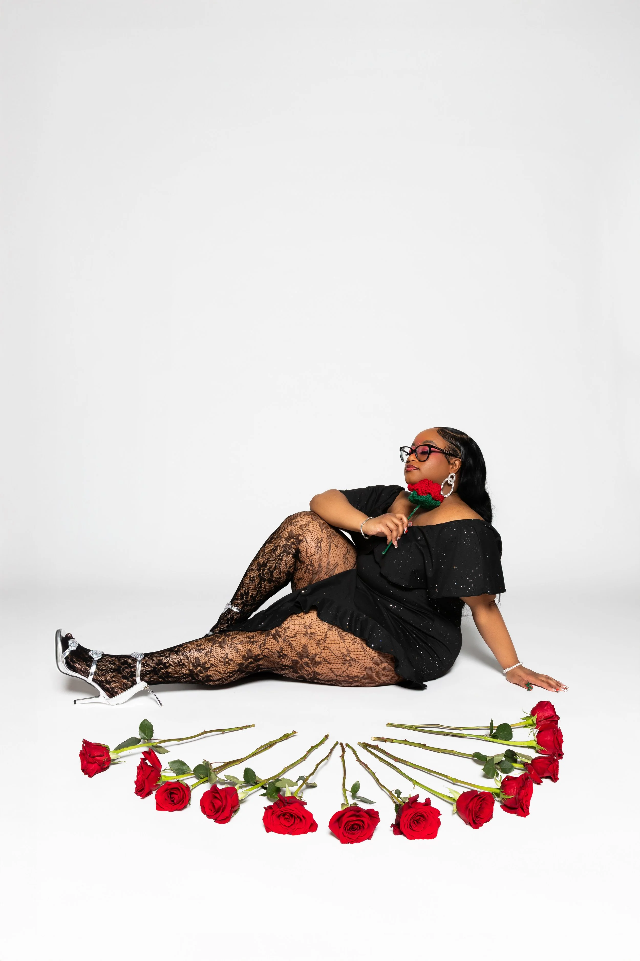 Woman wearing black dress with sparkles, lace stockings, silver heels, and glasses, posing on a white background while holding a red rose.