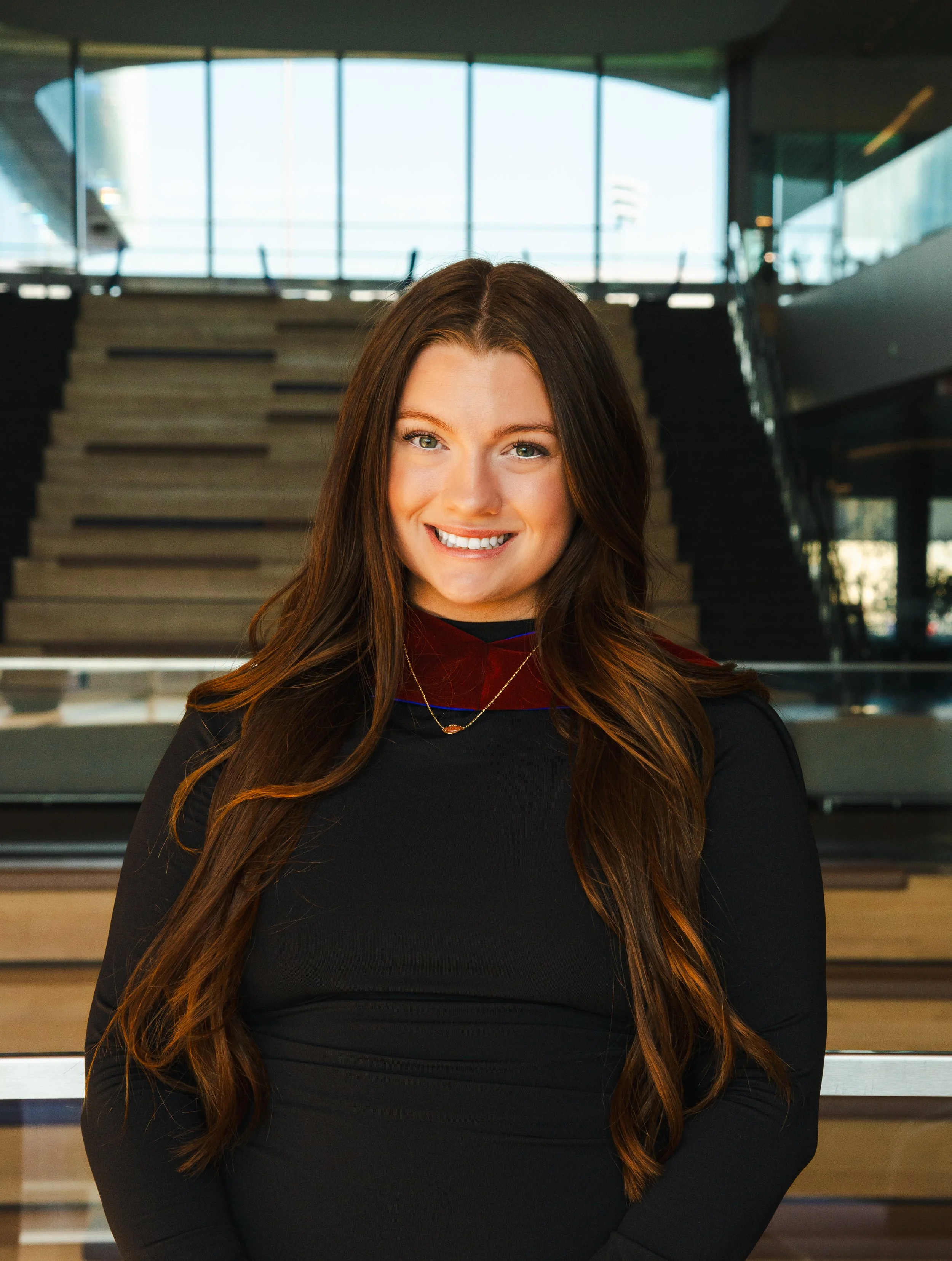 A young woman with long brown hair and blue eyes, smiling, wearing a black dress and a gold necklace, standing indoors with stairs and large windows in the background.