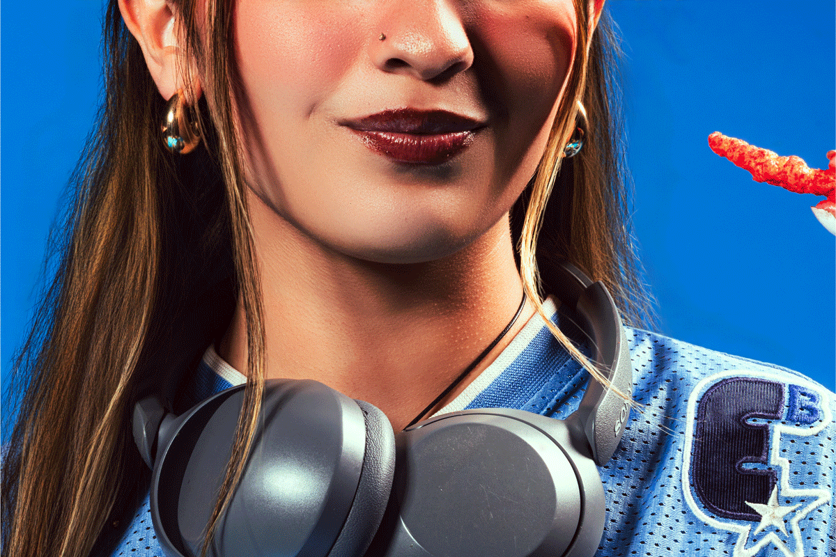 Close-up of a woman with long brown hair, earrings, wearing a blue sports jersey with a Dallas Cowboys star patch, and a pair of black headphones around her neck, against a blue background.