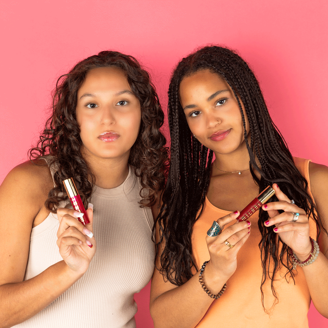 Two young women with curly hair holding lip gloss tubes against a pink background.