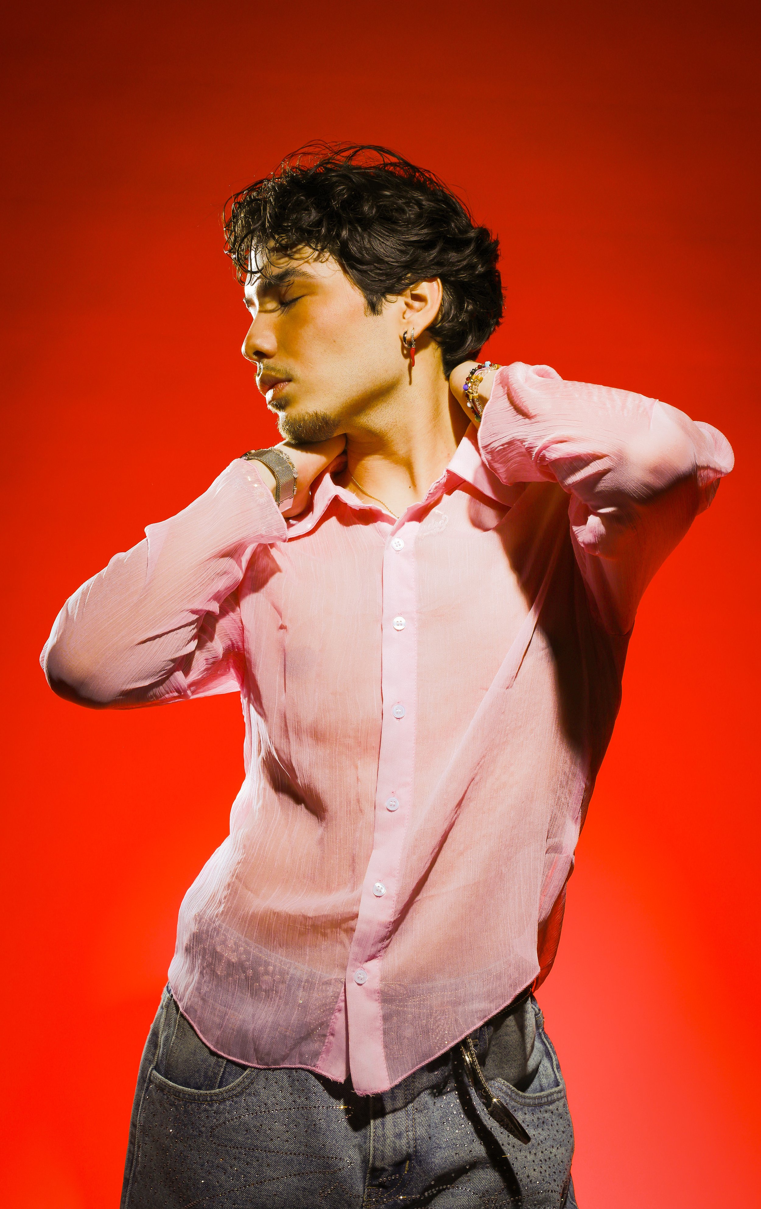 A young man with dark, curly hair and earrings, wearing a pink, slightly sheer button-up shirt and gray textured pants, striking a pose with his eyes closed against a red background.