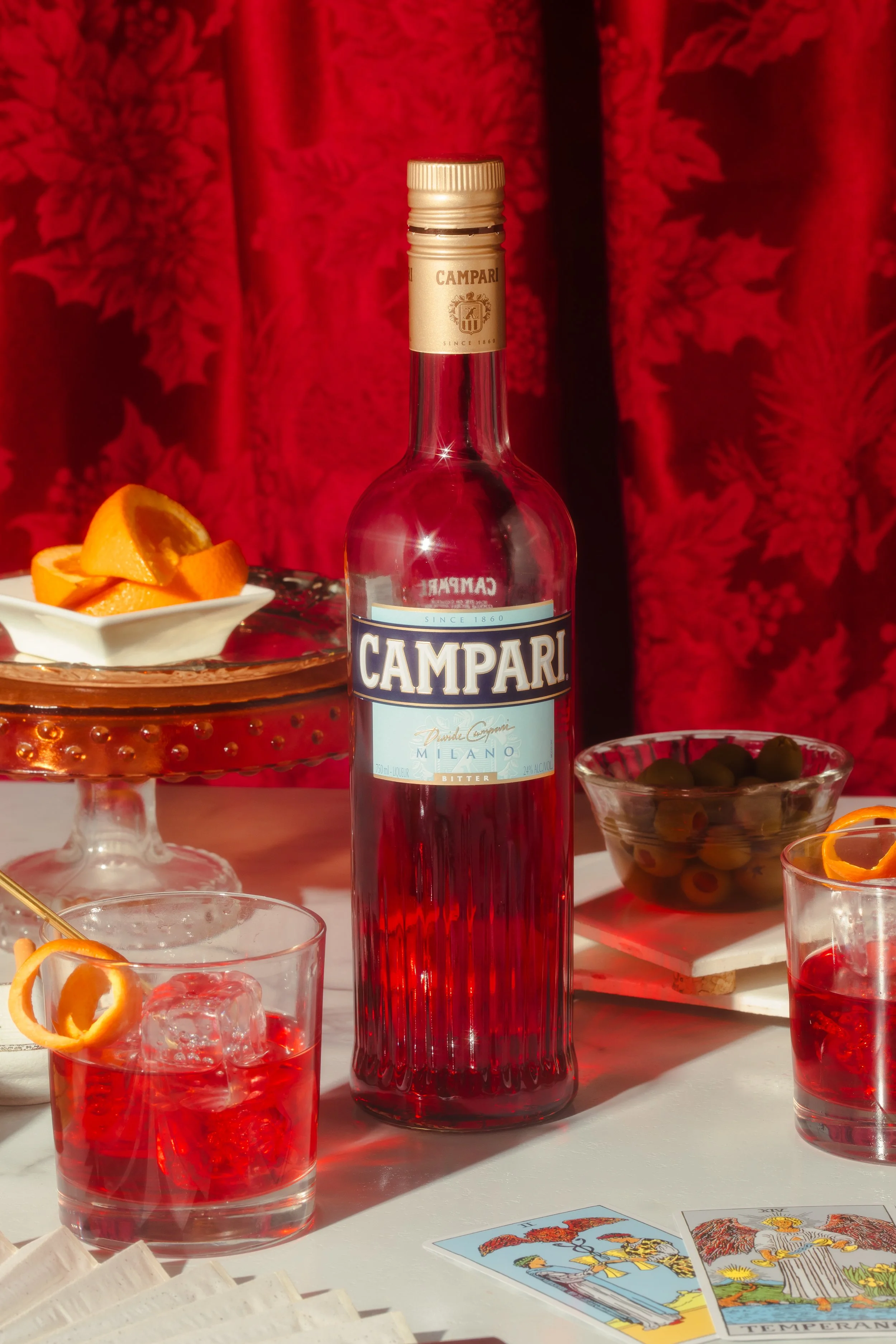A bottle of Campari aperitif surrounded by glasses of red-filled drinks, orange slices, and tarot cards on a white table with a red patterned curtain in the background.