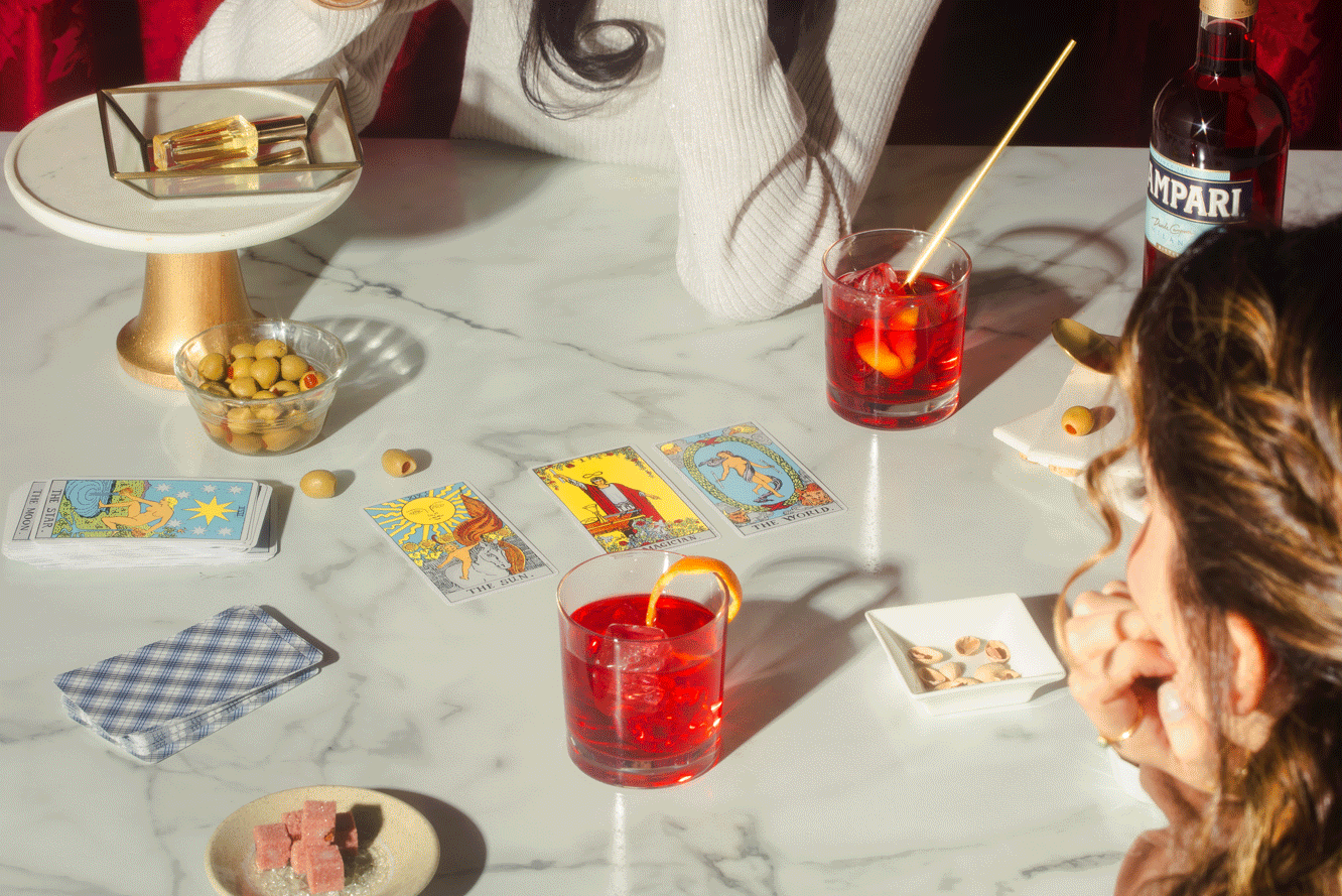 People playing tarot cards on a marble table, with drinks, snacks, and candies, in a cozy setting.