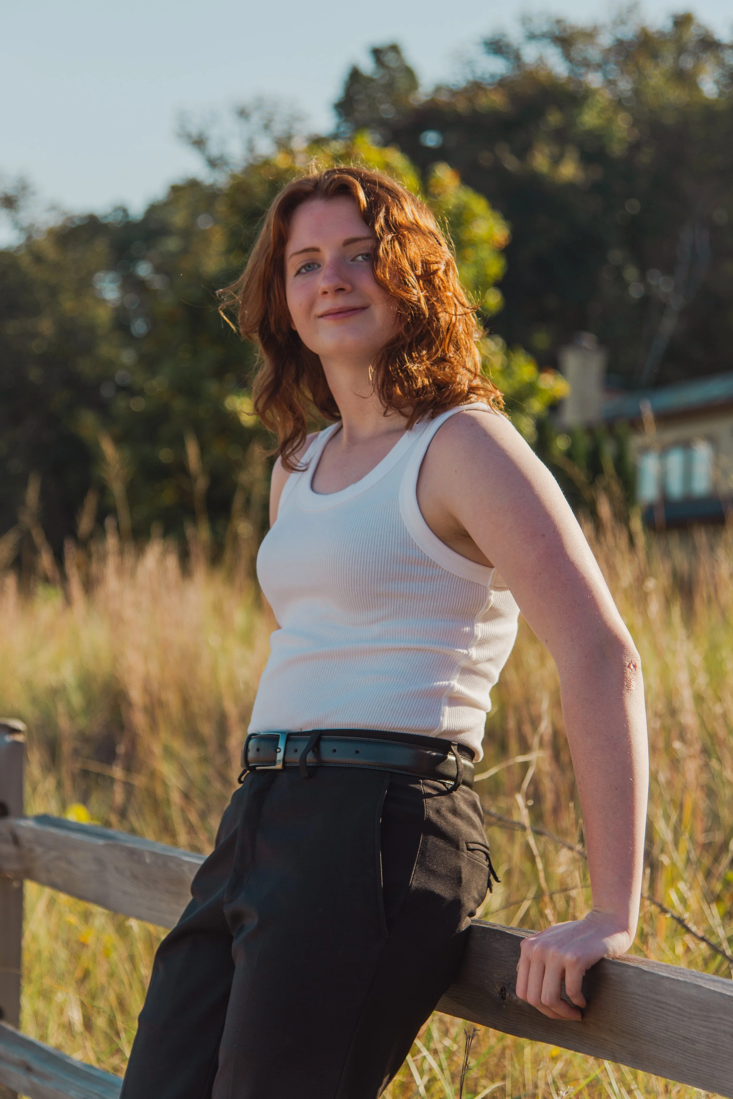 A young woman with red, wavy hair smiling and sitting on a wooden fence outdoors in a grassy field, wearing a white tank top and black pants, with trees and blue sky in the background.
