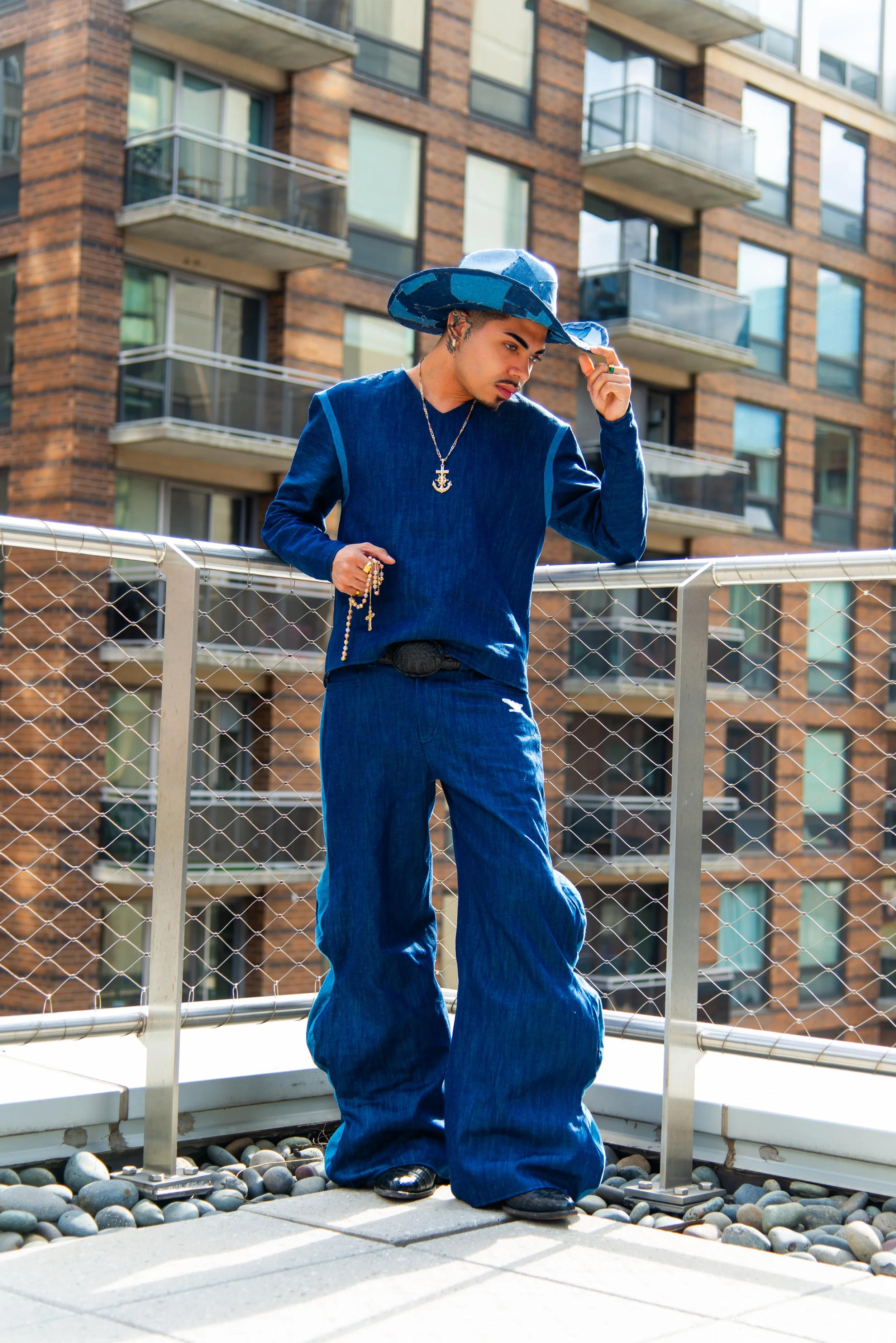 A young man in a blue wide-brimmed hat and blue denim clothing, wearing jewelry including rings, a necklace, and a rosary, stands on a balcony with a metal railing. Behind him is a multi-story brick building with glass balconies. The man is looking d