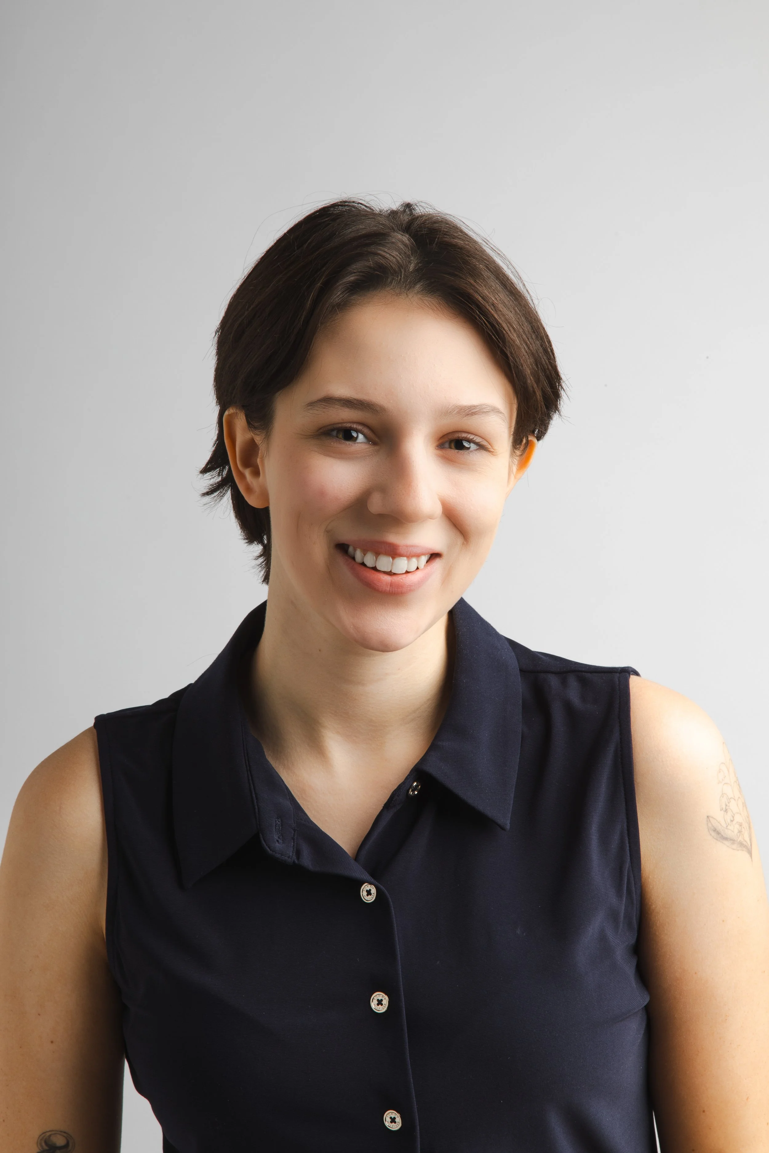 Close-up of a woman with short dark hair, wearing a sleeveless dark shirt, smiling against a plain light background.