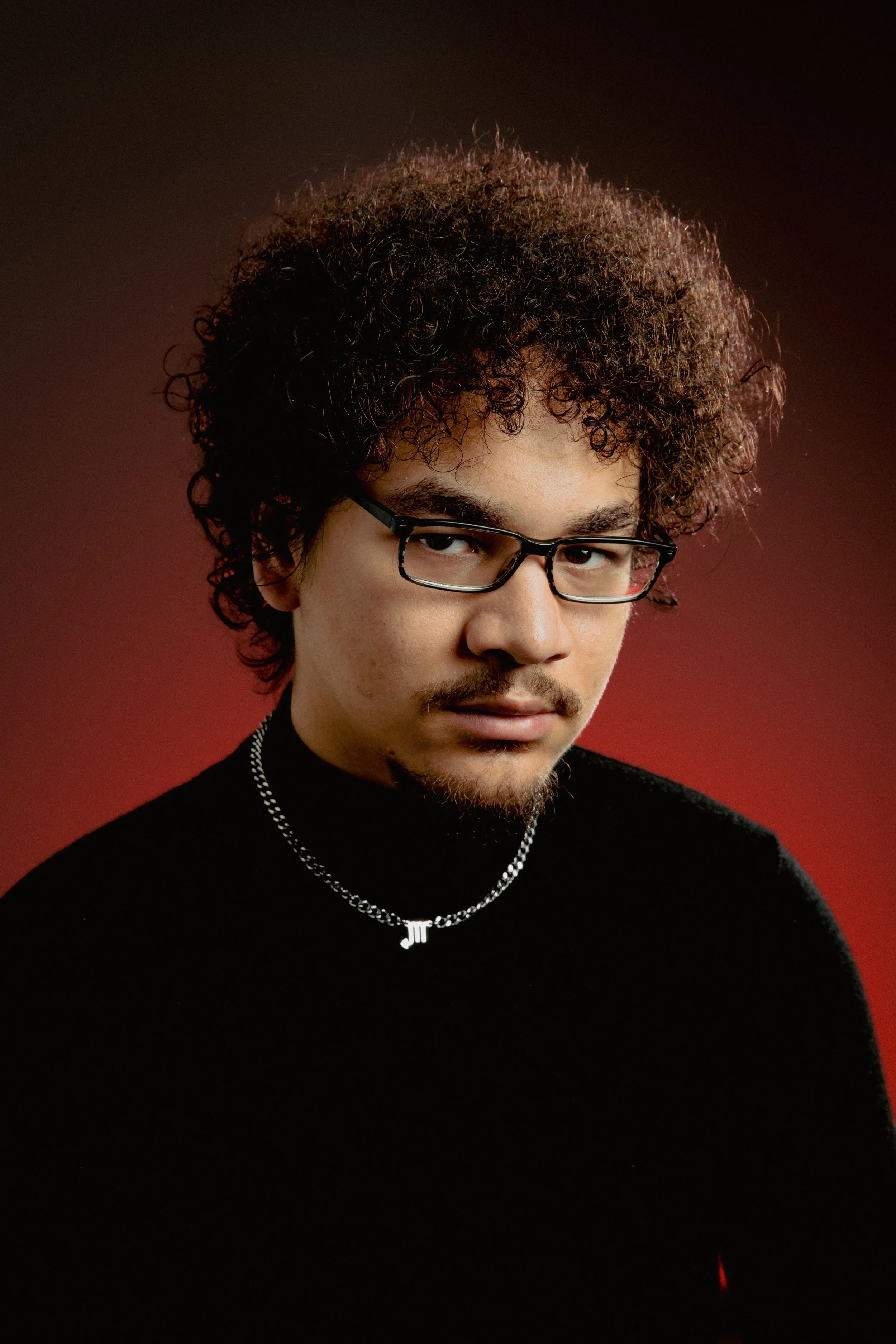 A young man with curly hair and glasses wearing a black turtleneck and a silver necklace with initials 'JT' against a red and black background.