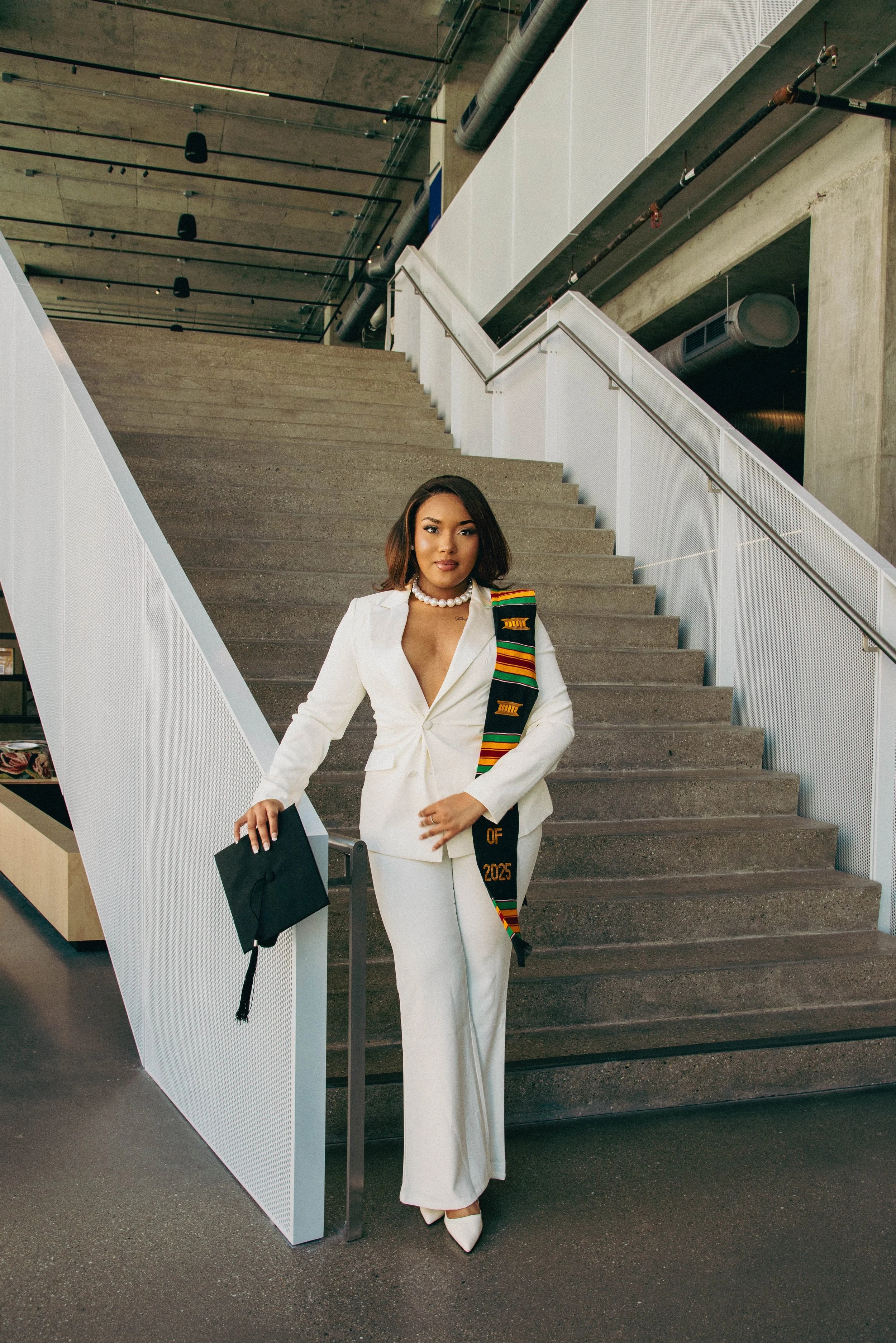 A woman dressed in a white suit with a pearl necklace, holding a black folder, standing on stairs in a modern, industrial-style building with concrete, metal, and exposed ductwork.