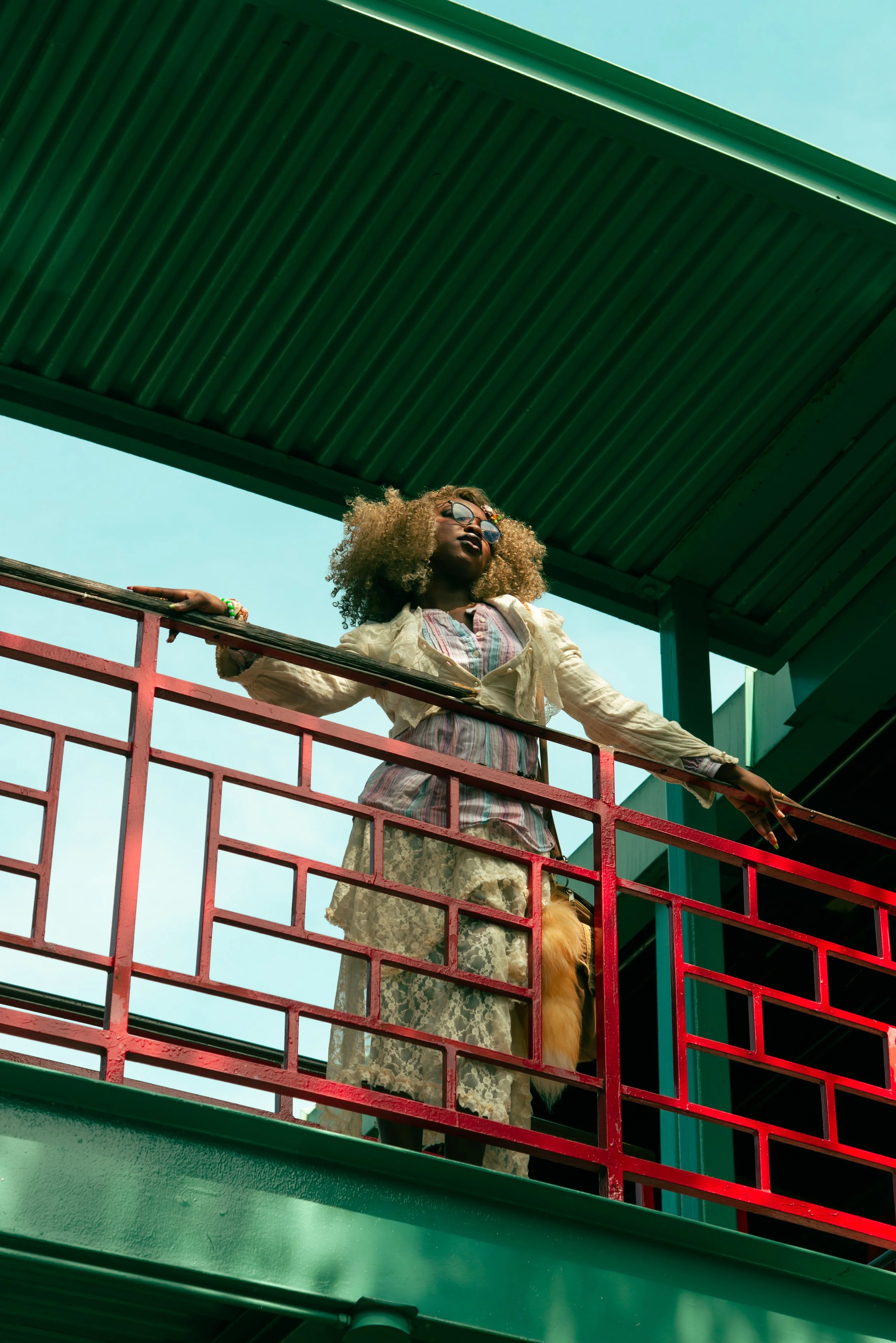 A woman with curly hair wearing glasses, a floral skirt, and a light jacket, standing on a colorful balcony with a red railing and green roof, looking into the distance.